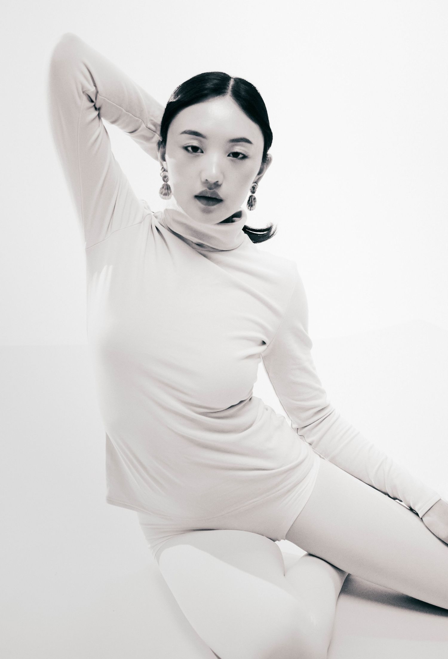 Black and white fashion portrait of a woman in a fitted turtleneck, seated with her arm raised behind her head