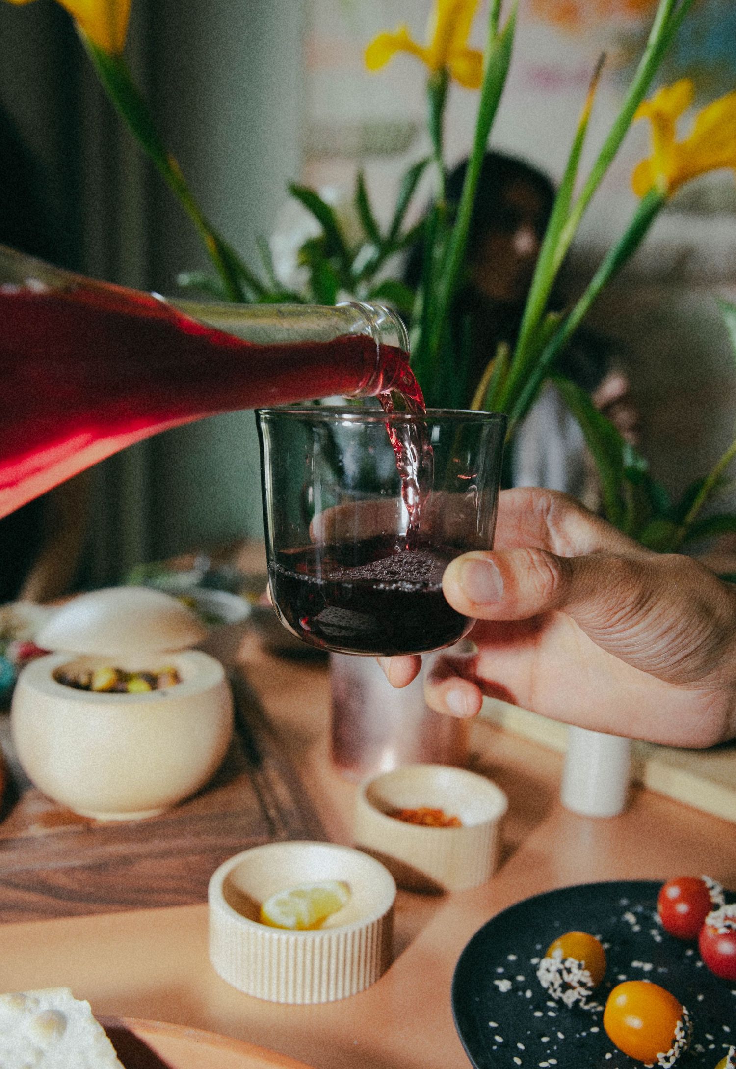 Red wine poured into a glass over a dinner table set with olives, small dishes, and yellow irises