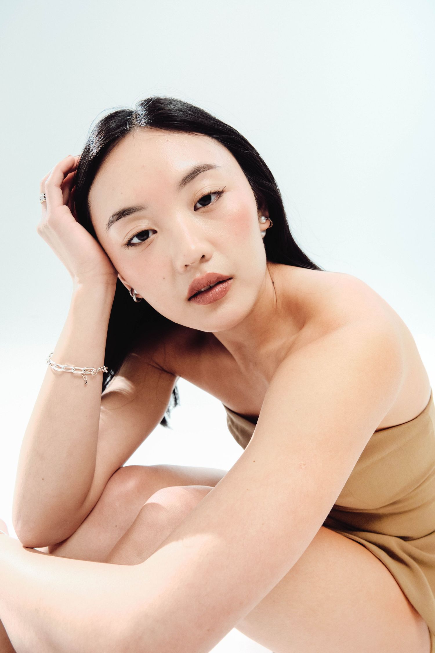 Studio portrait of a woman in a strapless taupe dress, resting her hand in her hair against a white background