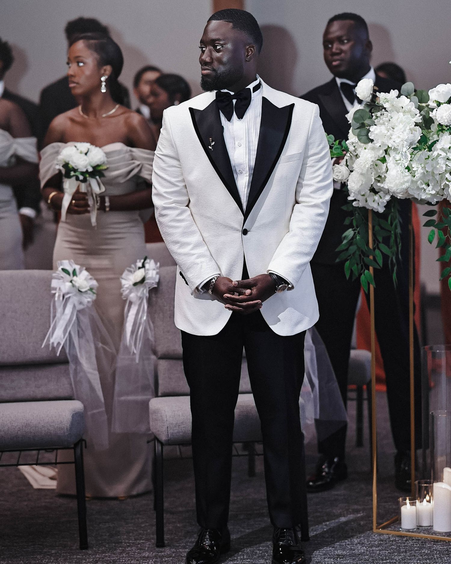 Groom in a white tuxedo and black bow tie standing at the wedding ceremony with bridesmaids and groomsmen behind him.