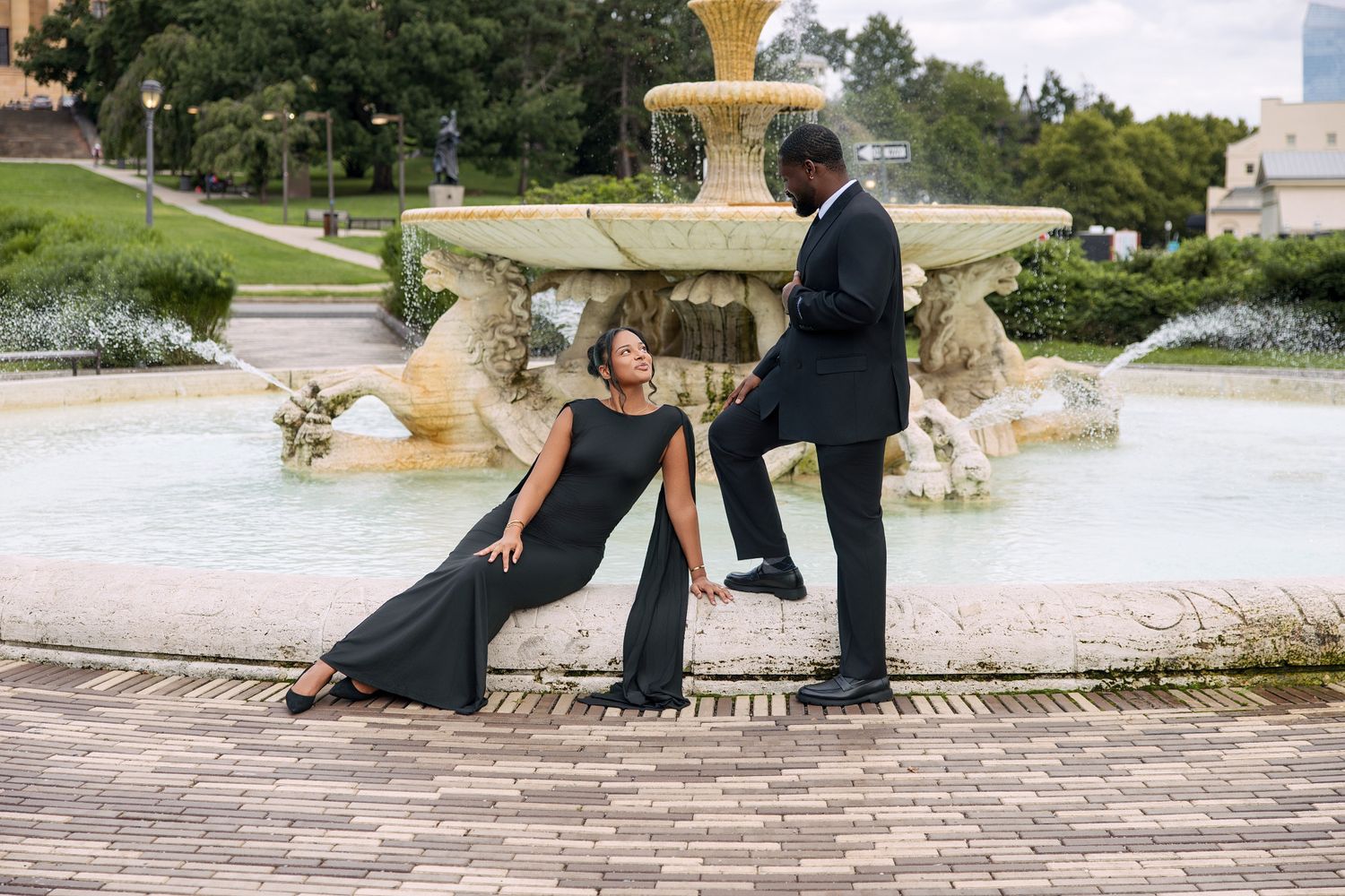 Couple dressed in elegant black outfits posing beside a decorative outdoor fountain.