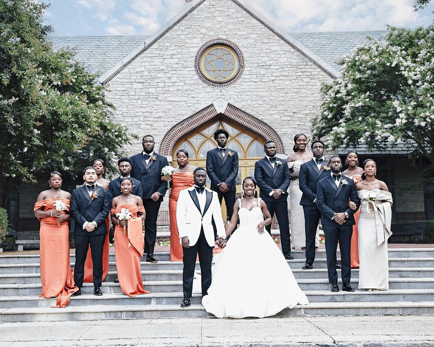 Bride and groom posing with bridesmaids in orange dresses and groomsmen in dark suits outside a church.