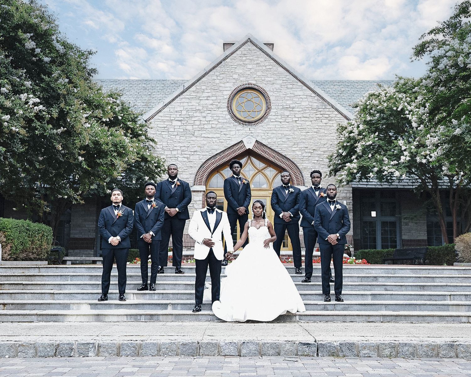 Bride and groom standing with groomsmen in black suits on the steps of a stone church.