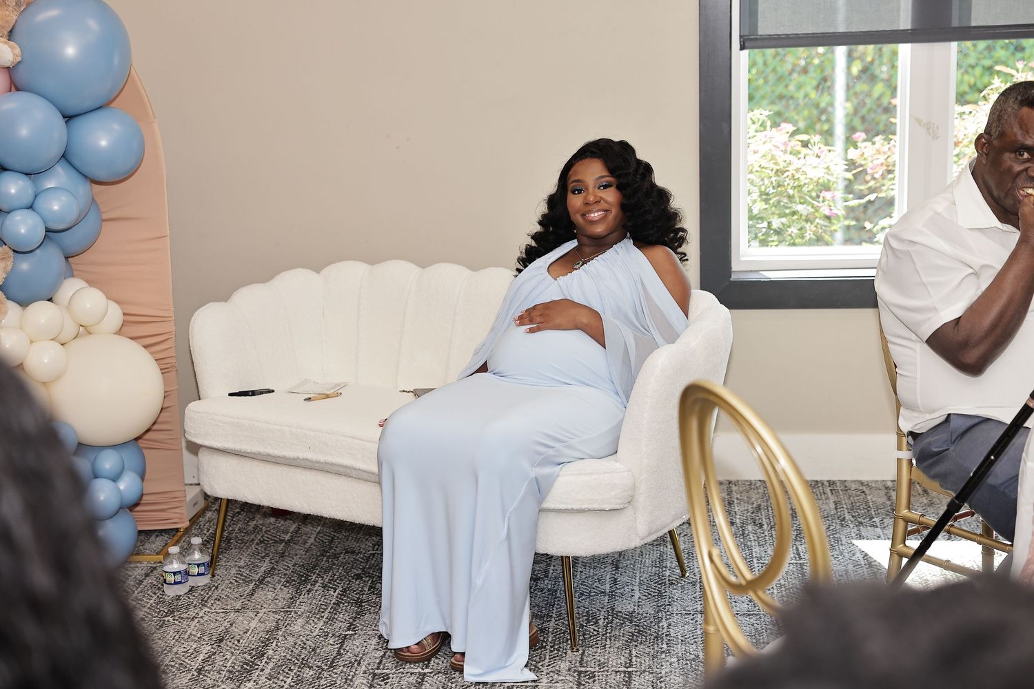 Expectant mother in a light-blue dress seated on a white couch at a baby shower celebration.