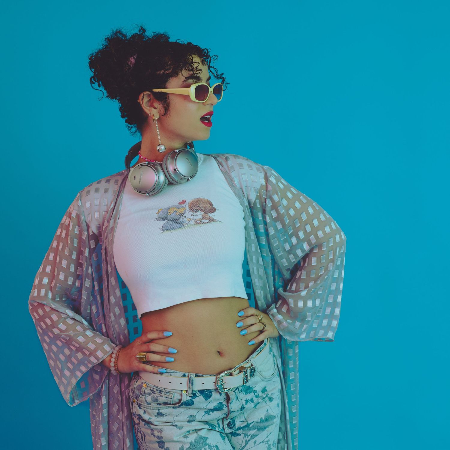 Stylish woman in crop top, patterned kimono, and sunglasses poses with headphones on blue background.