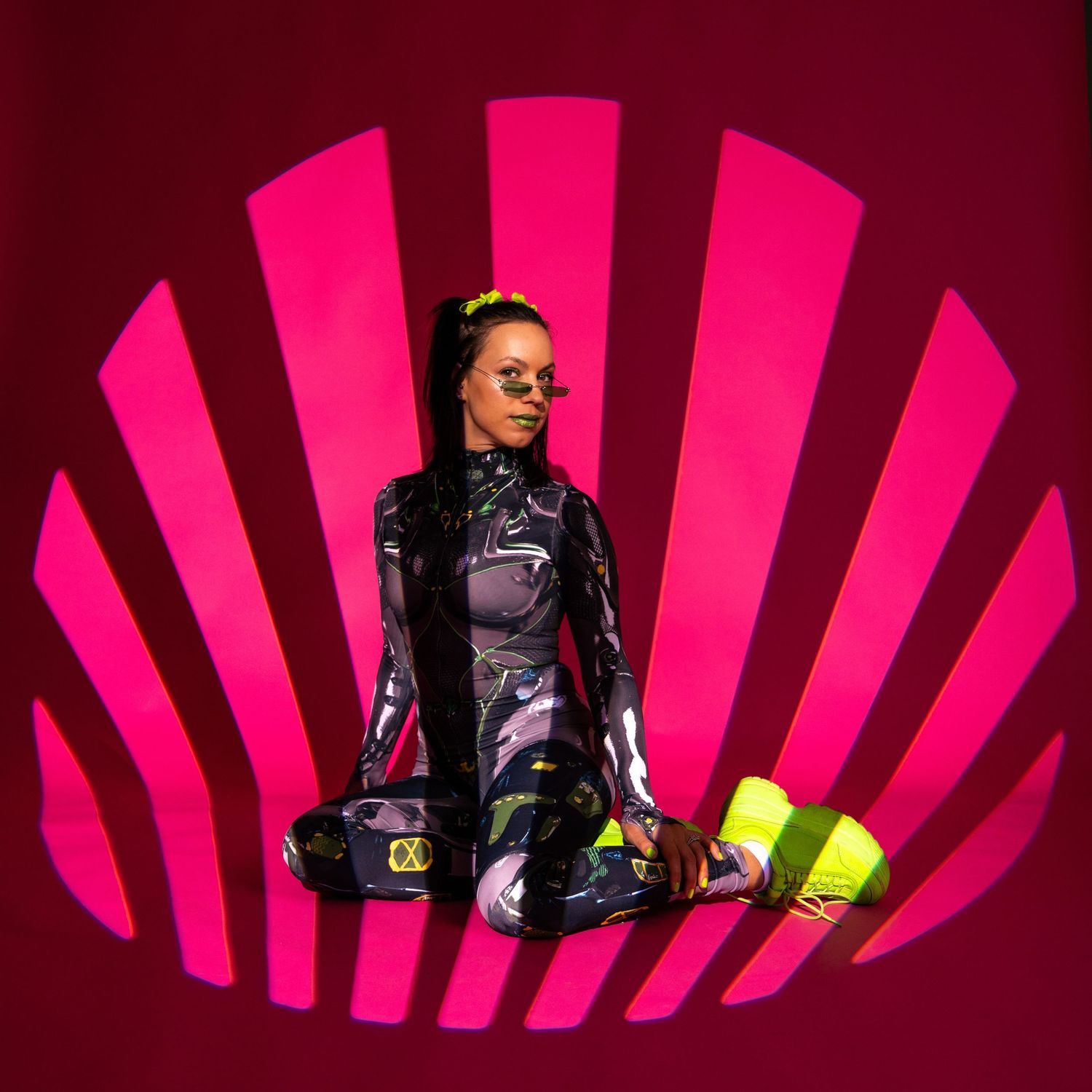 Woman in colorful bodysuit sits cross-legged in vibrant pink radial light installation.