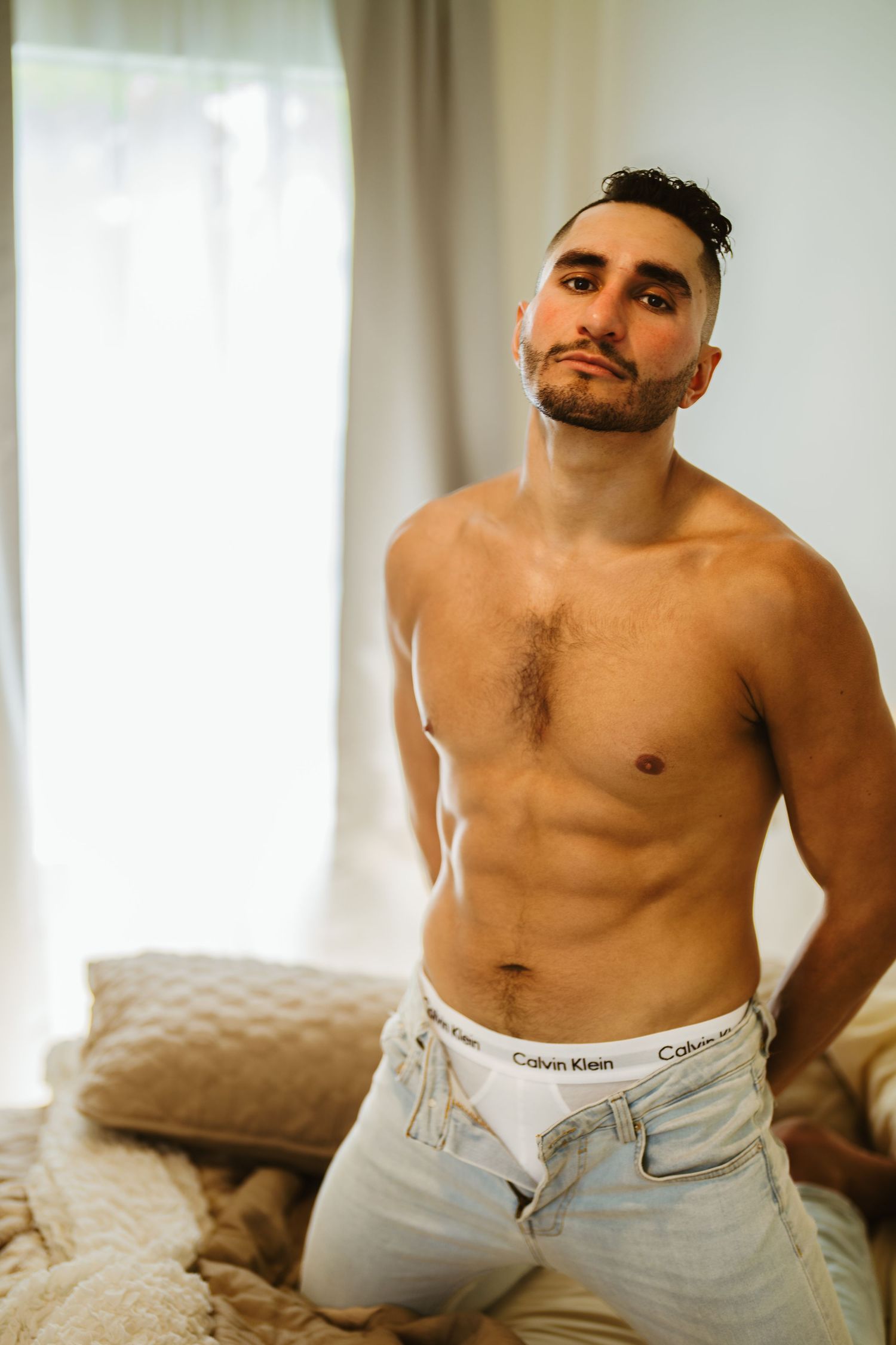 Shirtless bearded man in Calvin Klein jeans sits confidently on bed in bright room.