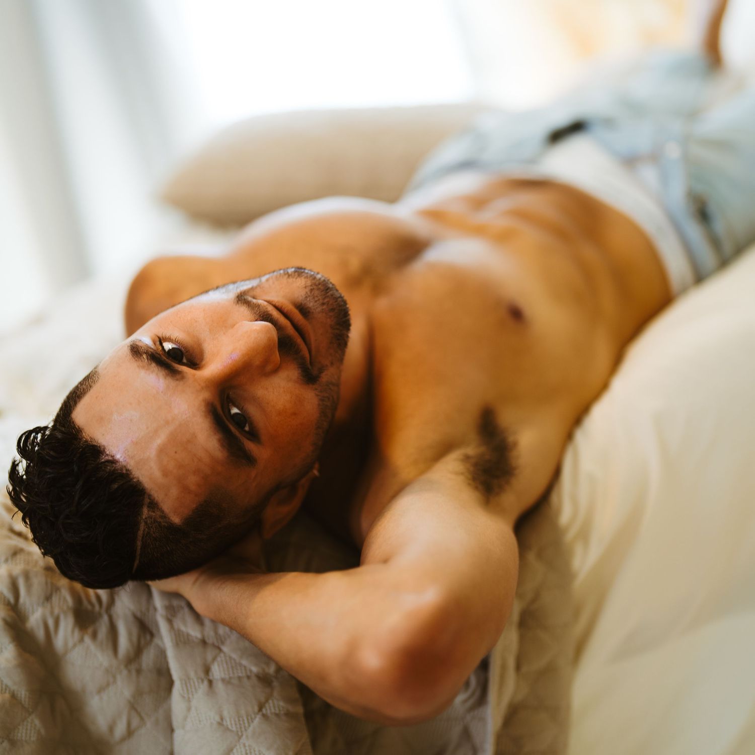 Shirtless athletic man with dark hair reclines on white bedding in natural window light.
