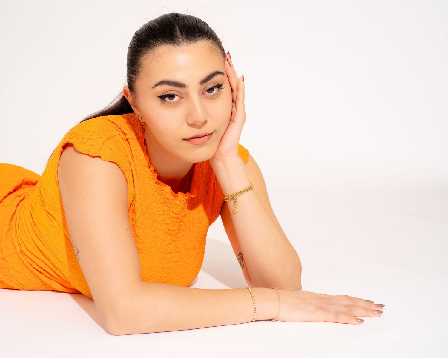 Woman in orange dress lying on white floor with hand on cheek in relaxed pose.