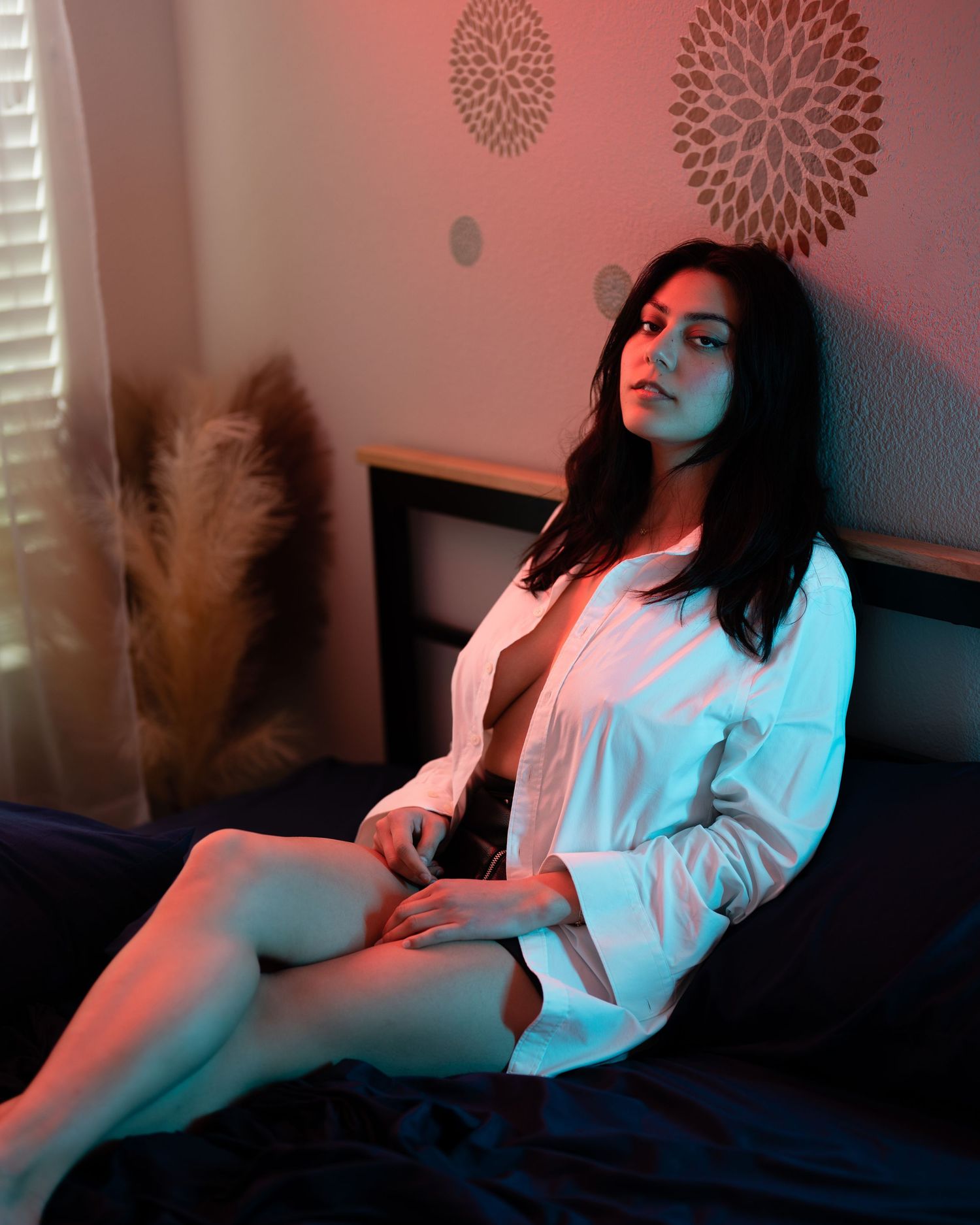 Woman in open white shirt seated on bed with dramatic red and blue neon lighting in bedroom.