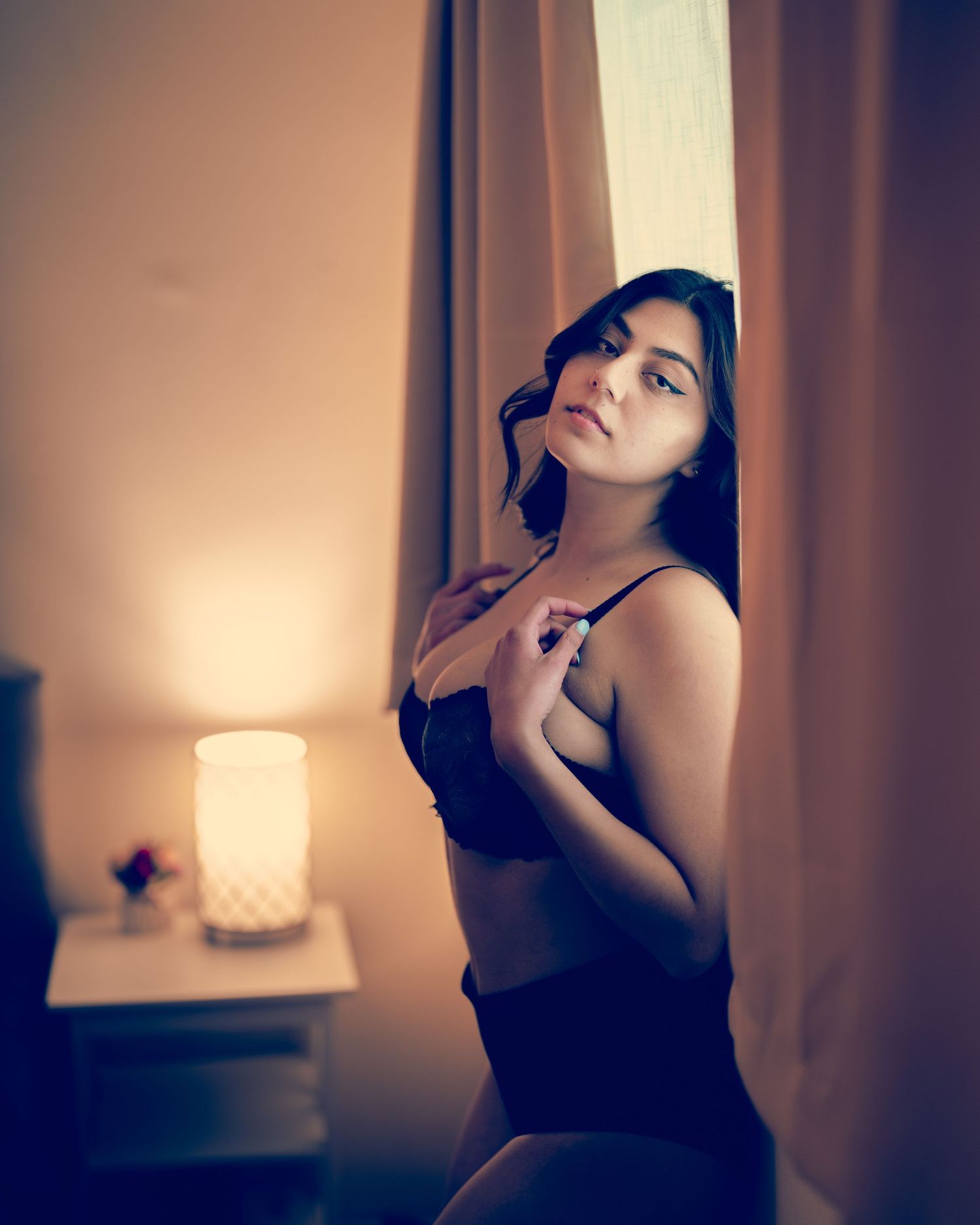 Woman in black lingerie standing by curtained window with warm lamp light in background.