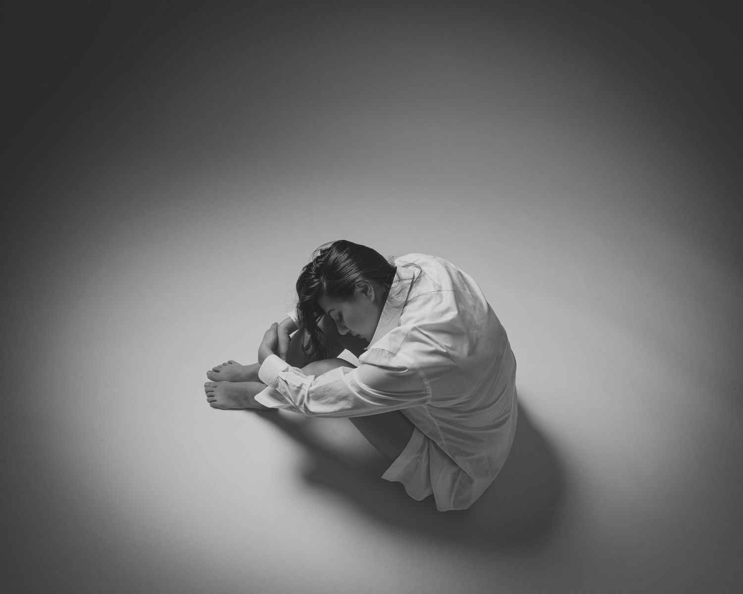 Black and white photo of person curled up on floor in white shirt, head down in contemplative pose.