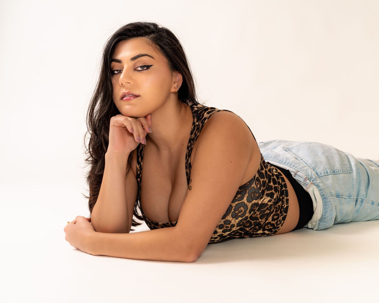Young woman in leopard print crop top and jeans lying on white floor, resting chin on hand.
