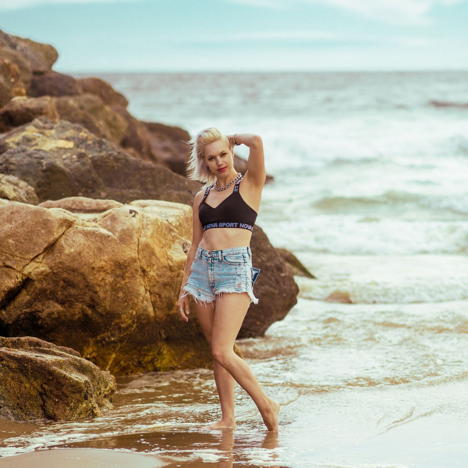 Blonde woman in black crop top and denim shorts stands barefoot on rocky ocean shoreline.
