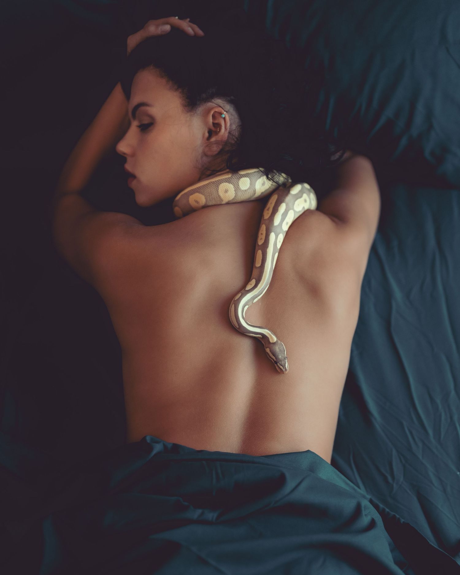 Artistic portrait of woman lying down with a ball python snake draped across her back.