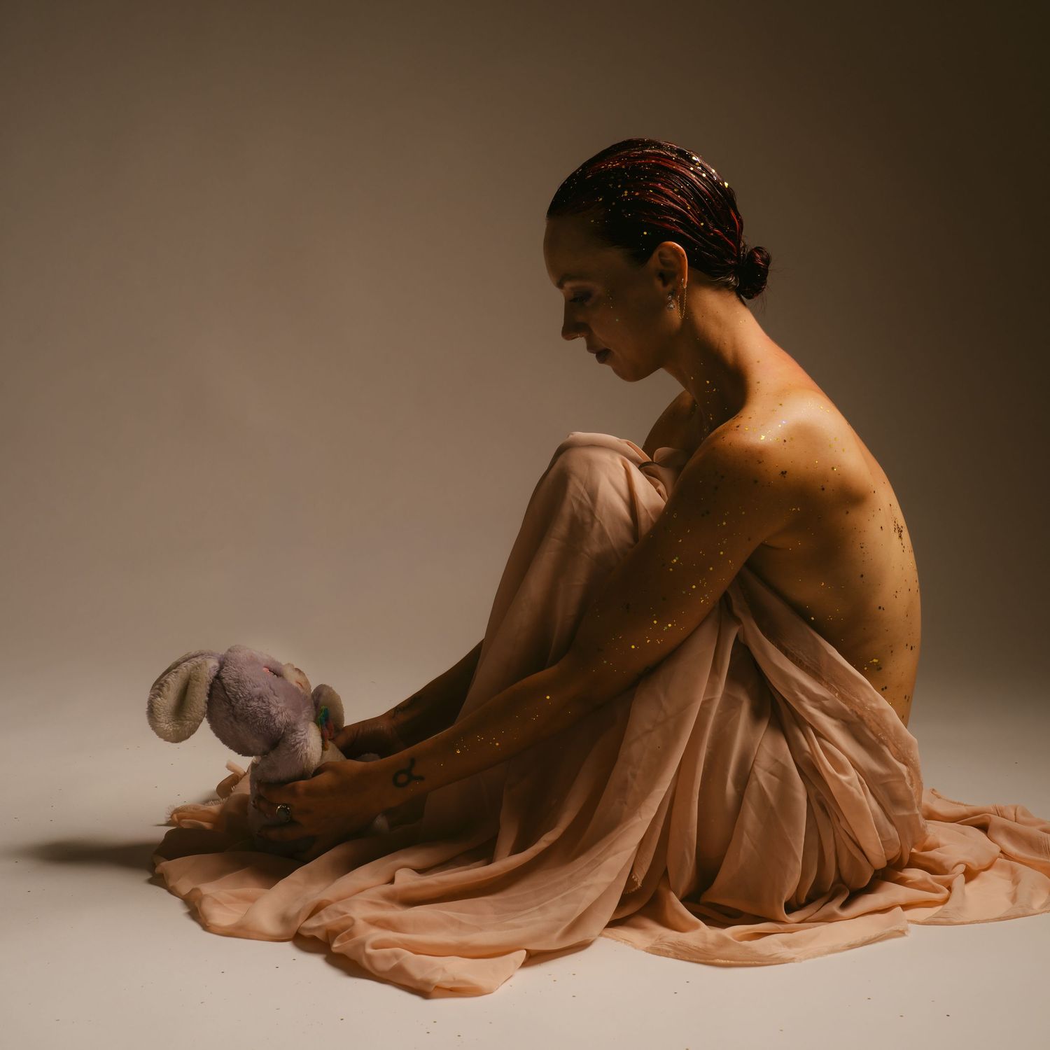 Woman wrapped in sheer fabric sitting with stuffed bunny in studio.