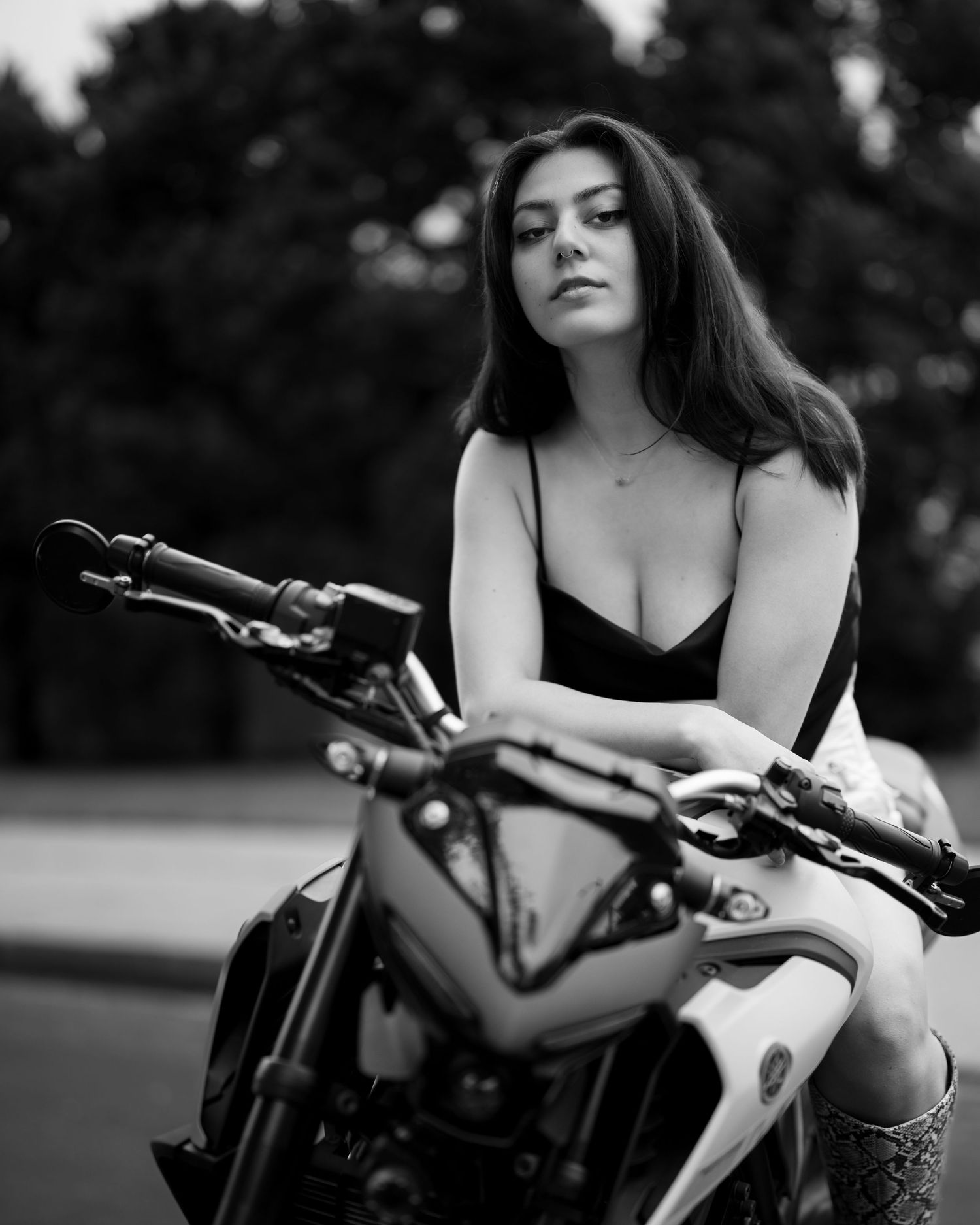 Black and white portrait of woman in black dress seated on motorcycle outdoors among trees.
