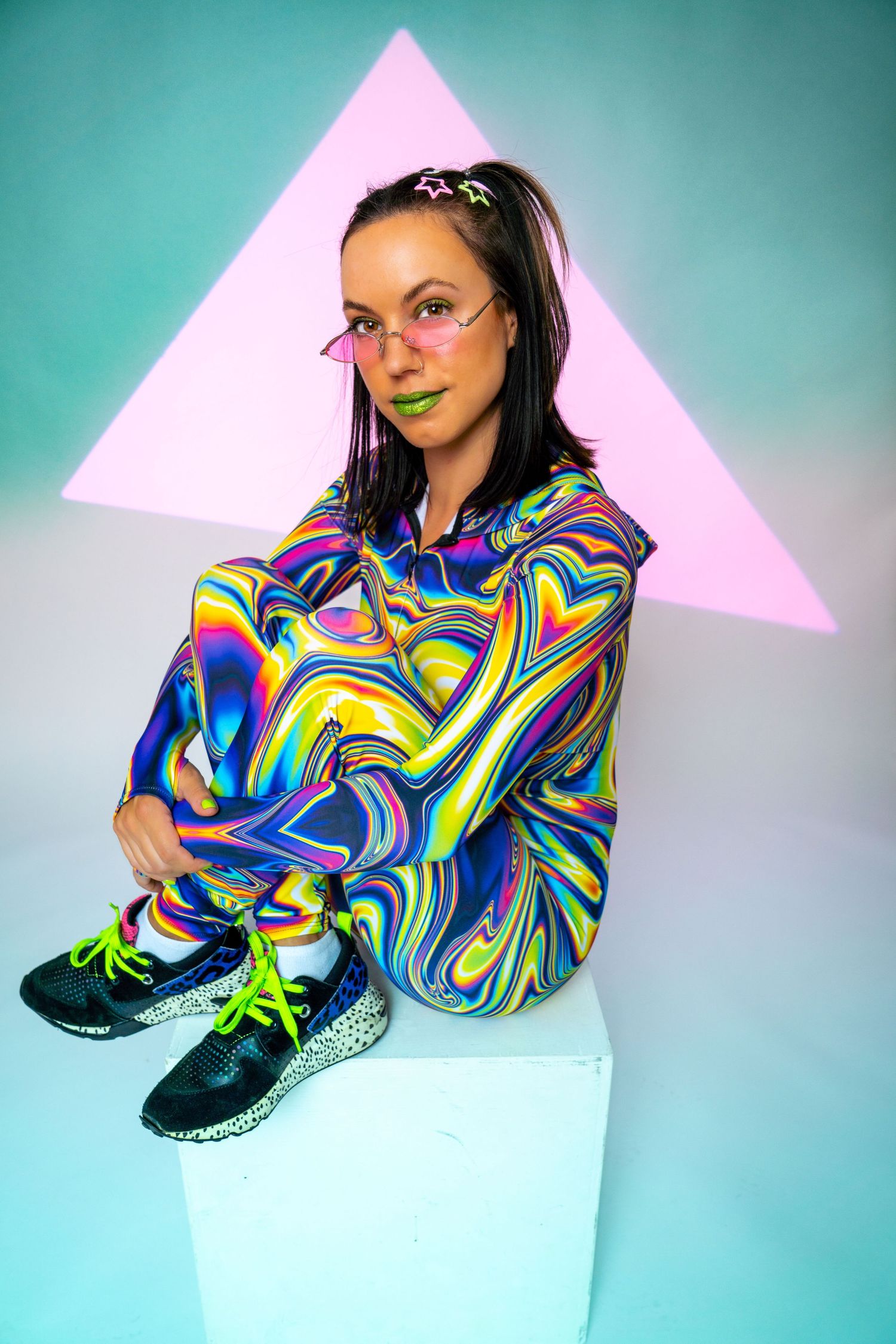 Woman in rainbow holographic tracksuit sits on white cube before teal background with pink triangle.