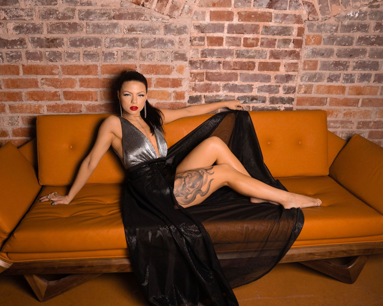 Elegant woman in black satin gown posing on orange sofa against brick wall backdrop.