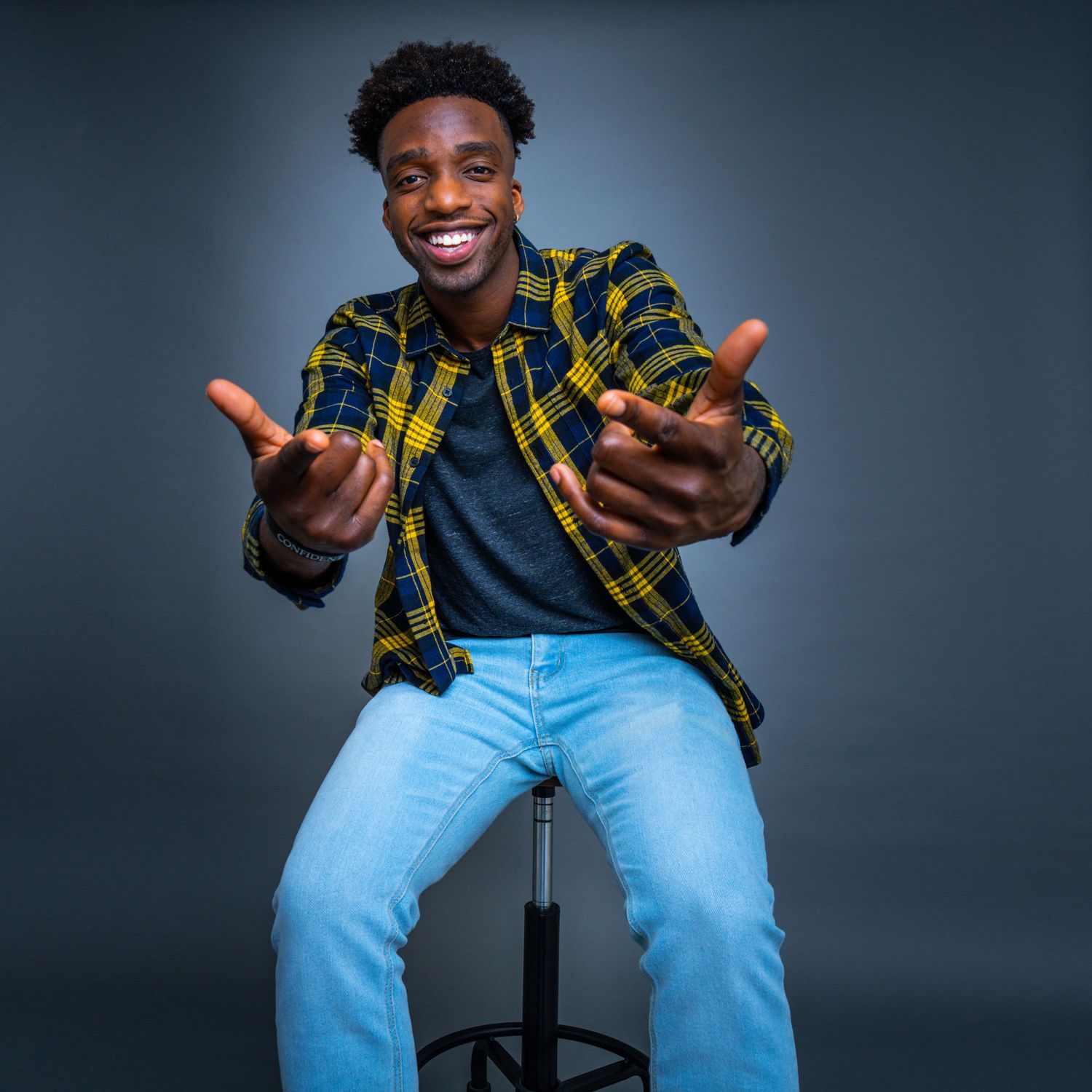 Smiling young Black man in plaid shirt and jeans sits on stool gesturing toward camera.