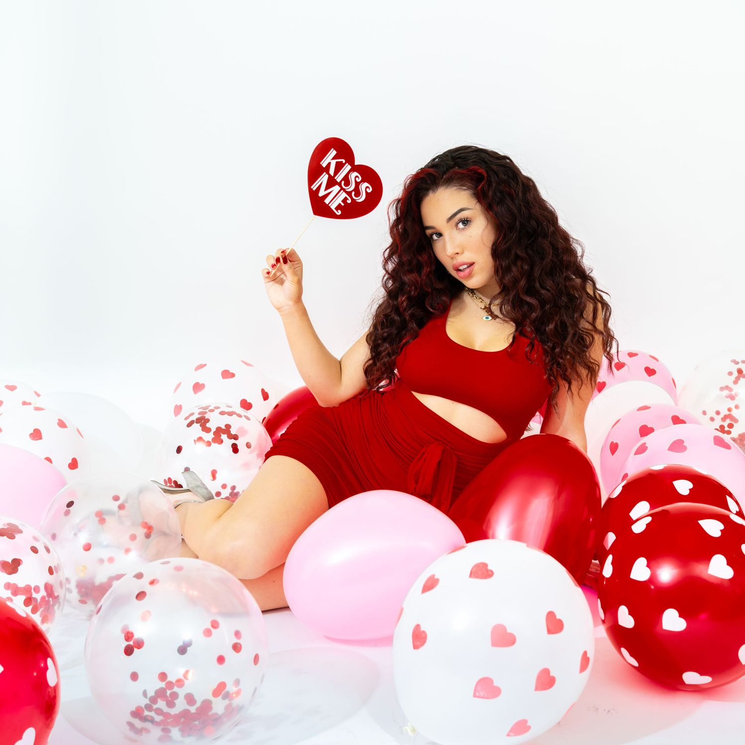 Woman in red outfit sitting among Valentine's Day balloons holding kiss me sign.