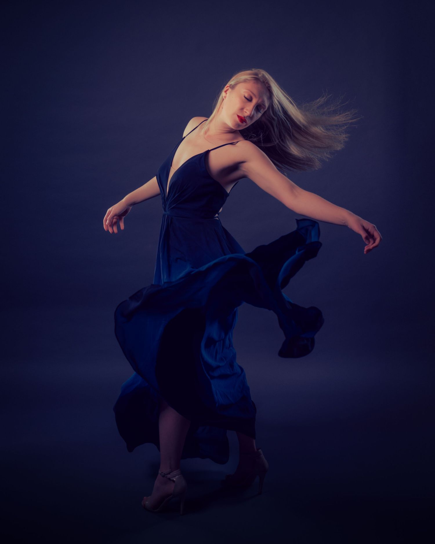 Blonde dancer in flowing blue gown leaps dramatically against dark blue studio background.