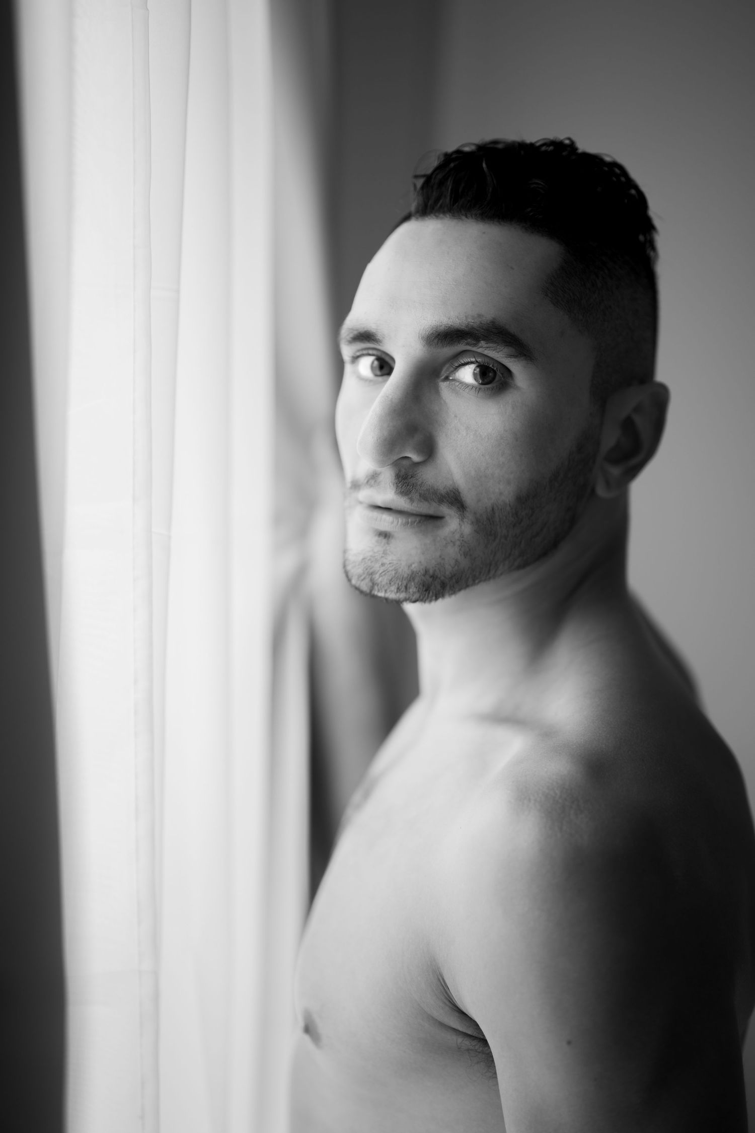 Black and white portrait of a shirtless man with dark hair standing by a window.