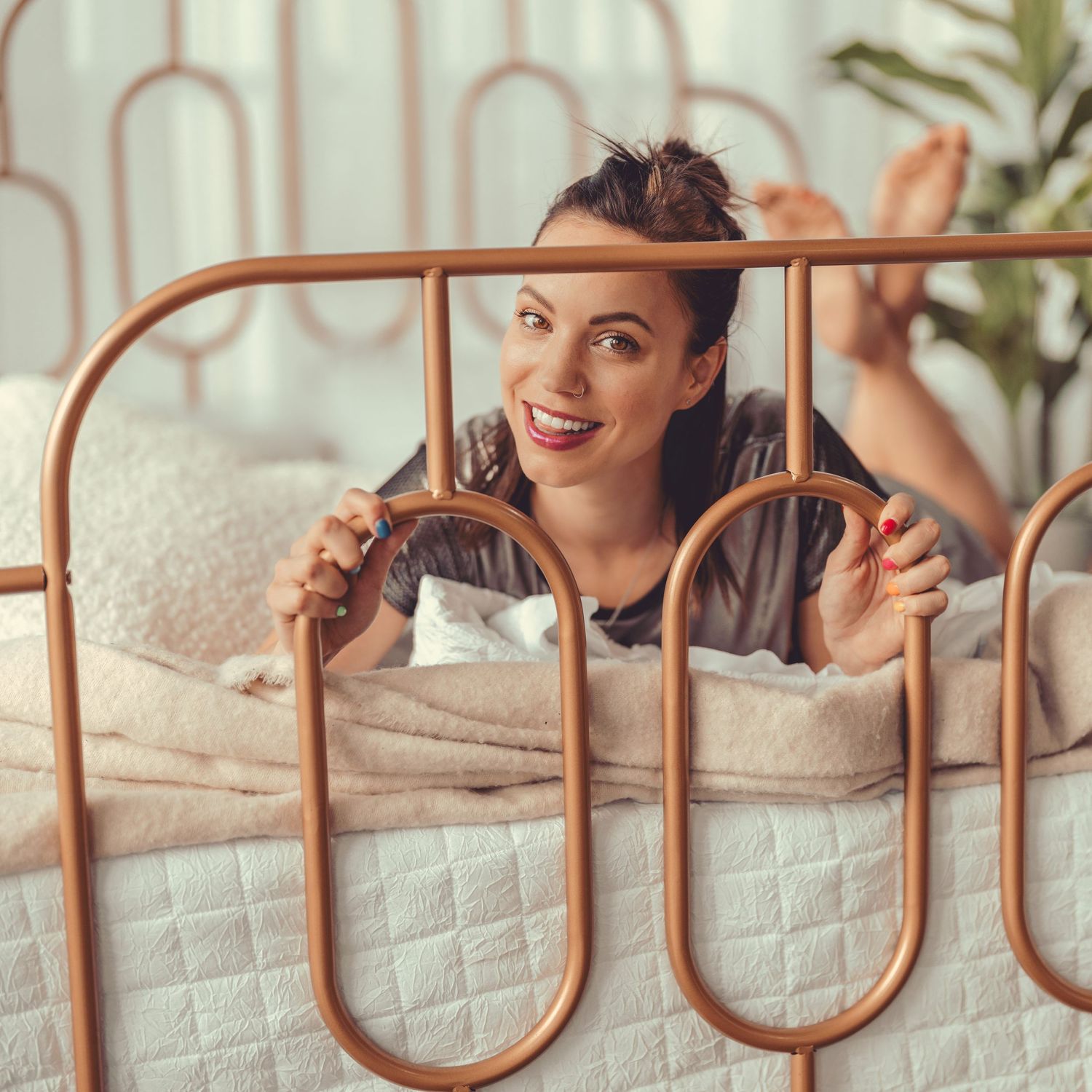 Smiling woman lying on bed peering over gold metal headboard.