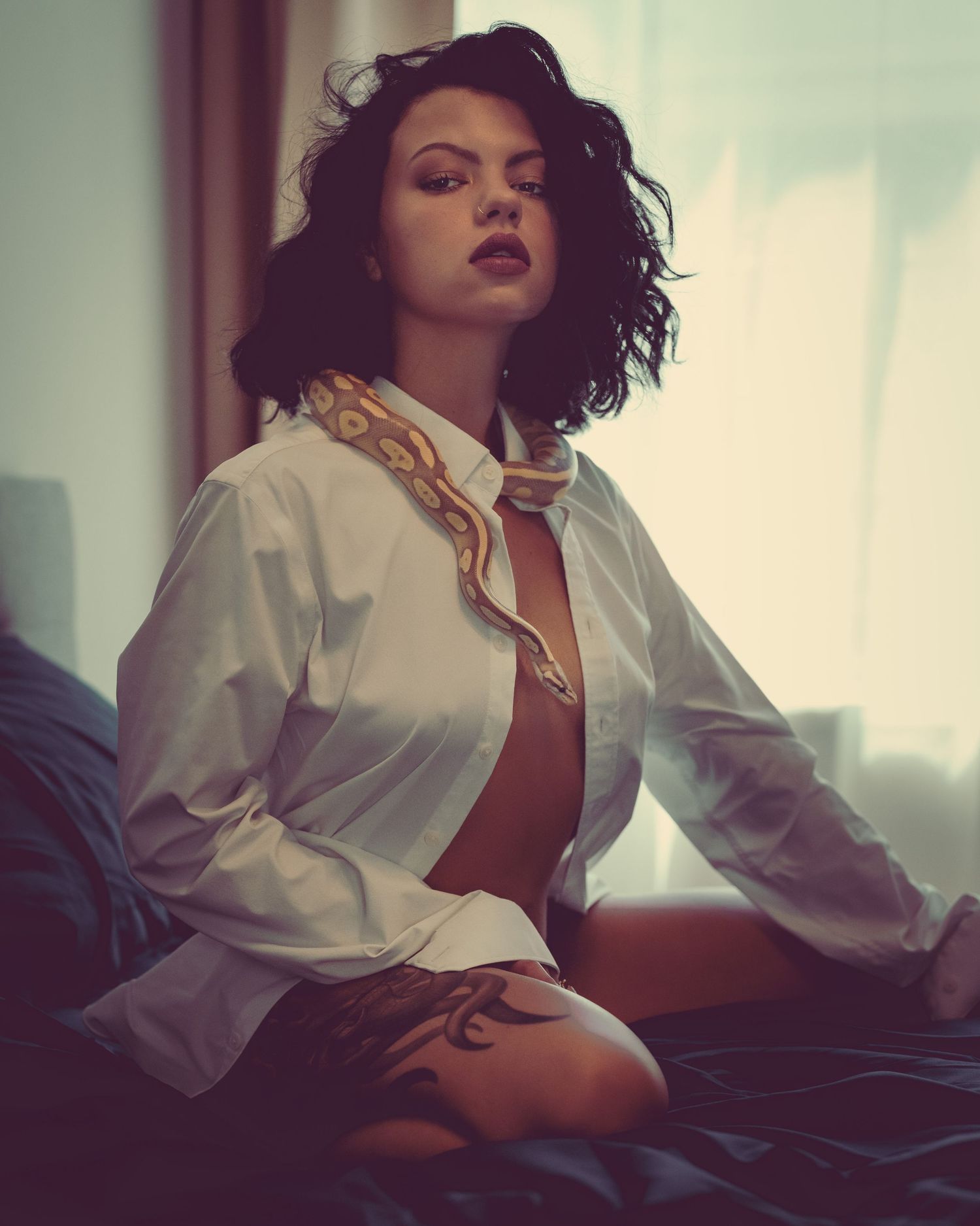 Young woman in open white shirt with snake draped around neck, seated on bed with tattoos visible.