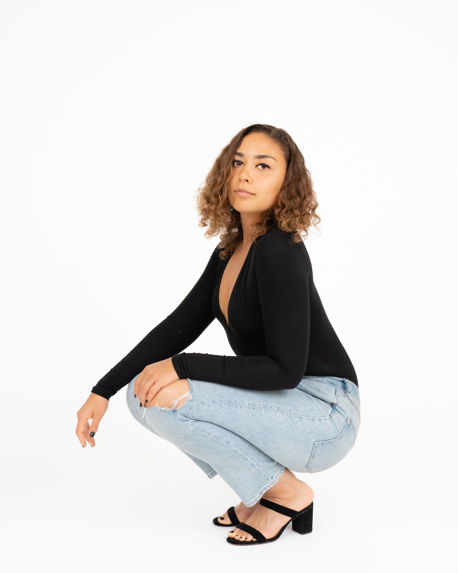 Woman in black bodysuit and light jeans crouching on white background.