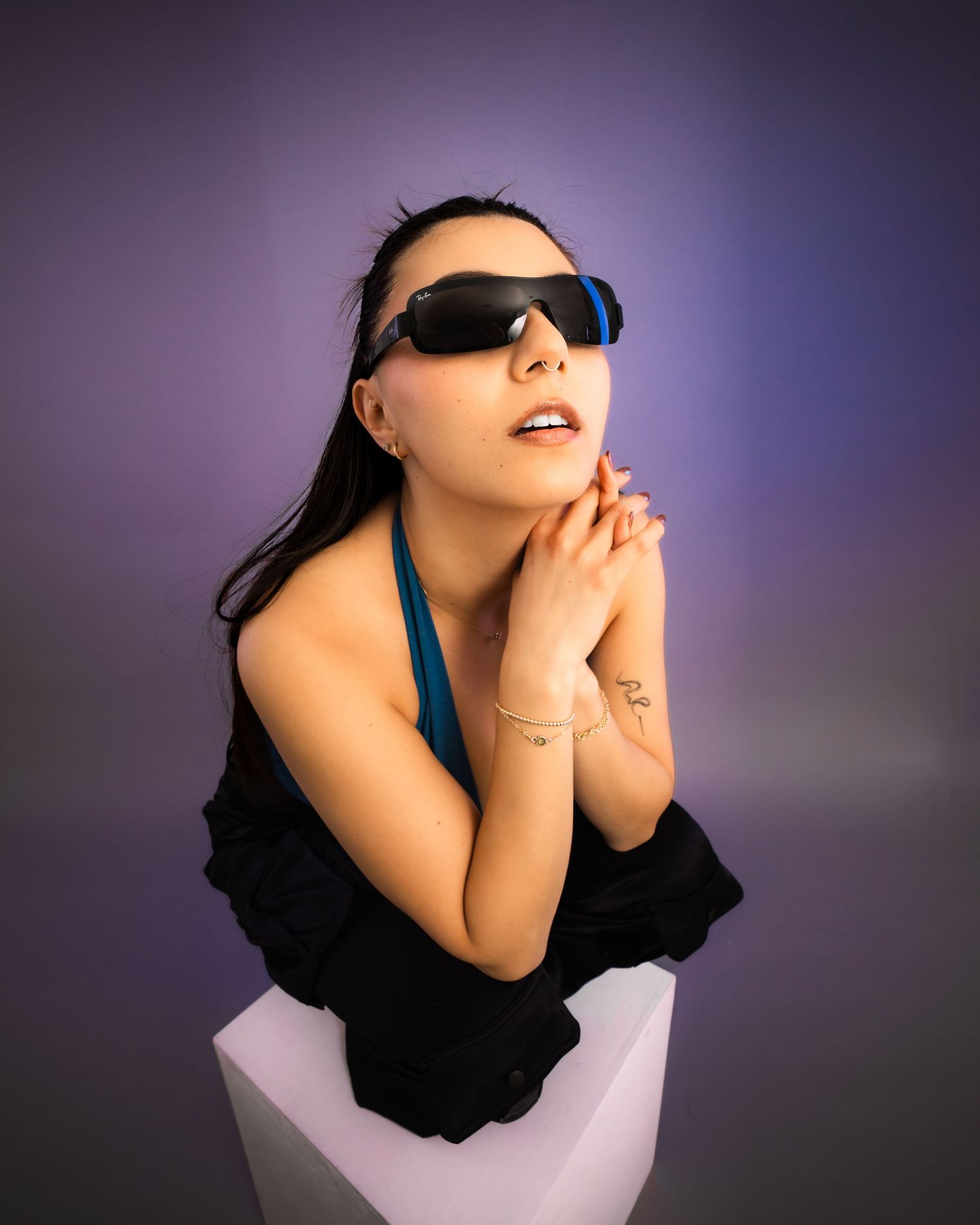Stylish woman wearing black sunglasses sitting on white pedestal against purple background.