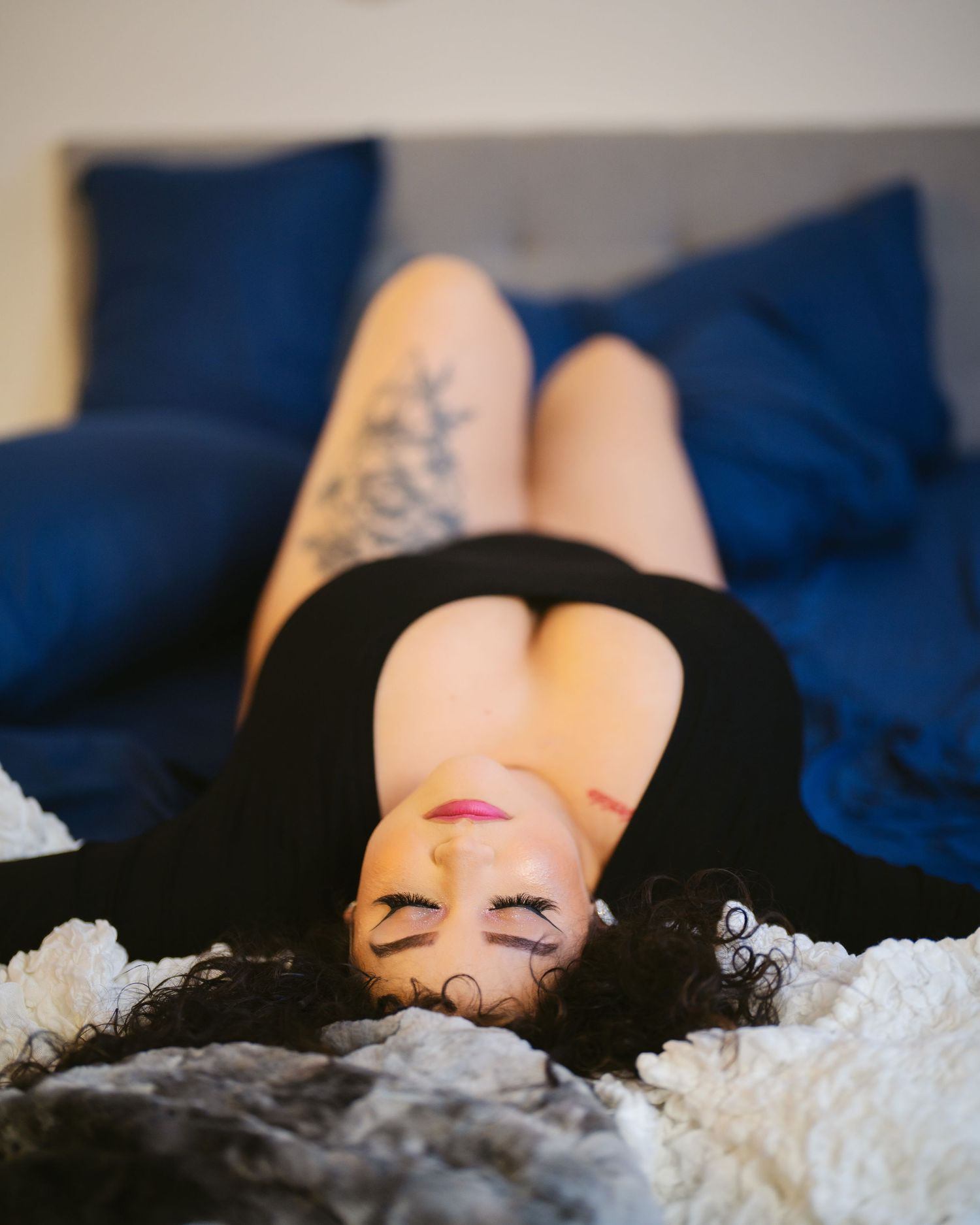 A woman with tattoos and dark curly hair lies on a bed with blue pillows, wearing a black dress.