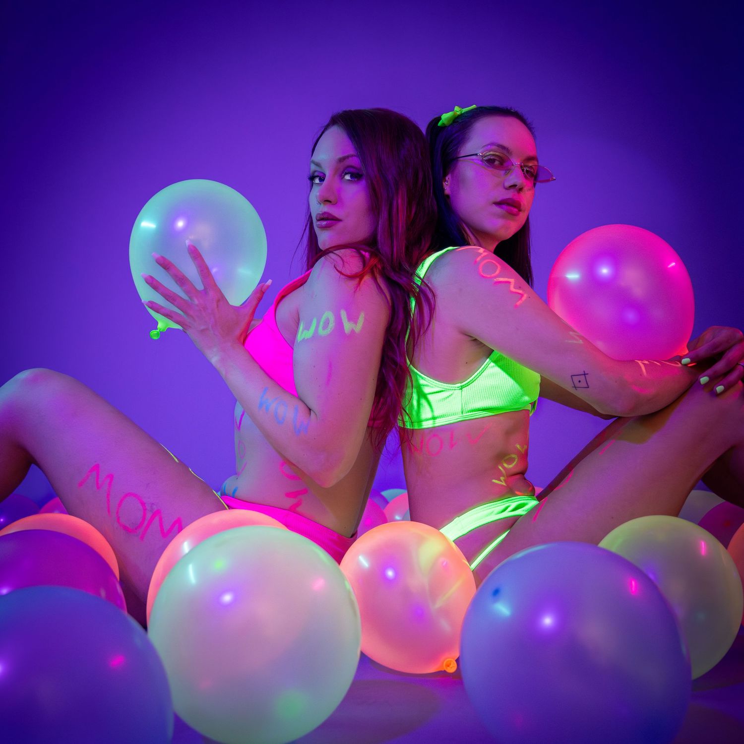 Two women in neon bikinis pose back-to-back surrounded by glowing balloons on purple background.