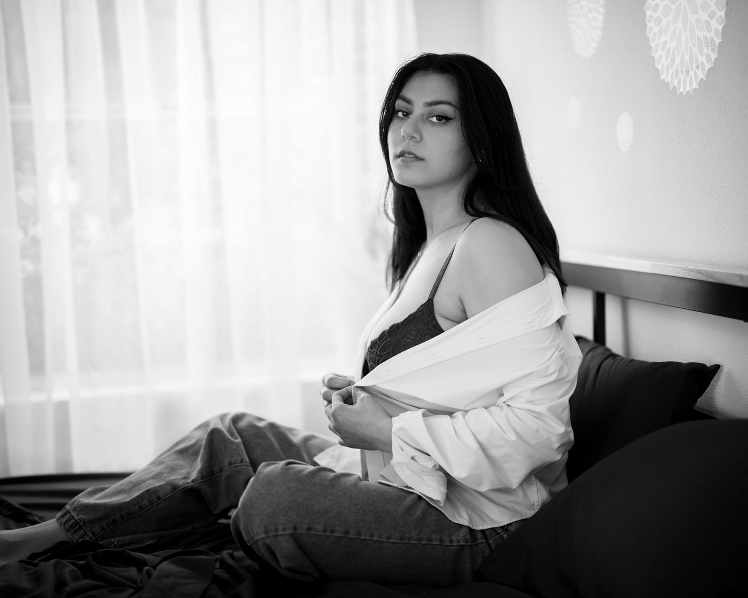 Black and white photo of woman in open white shirt and jeans sitting on bed by window.