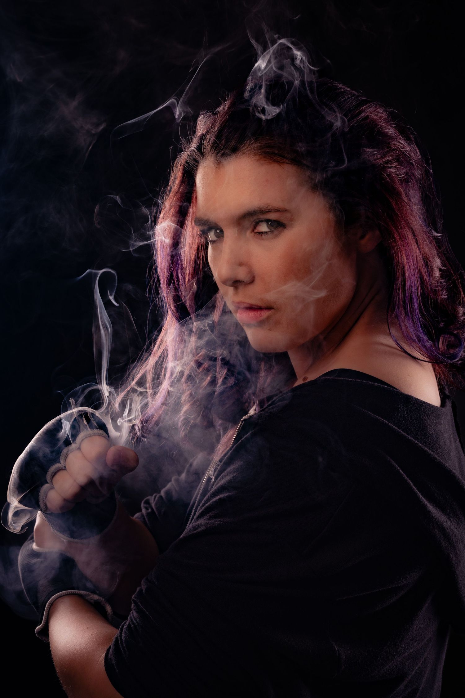 Woman with colorful hair poses dramatically surrounded by swirling smoke on dark background.