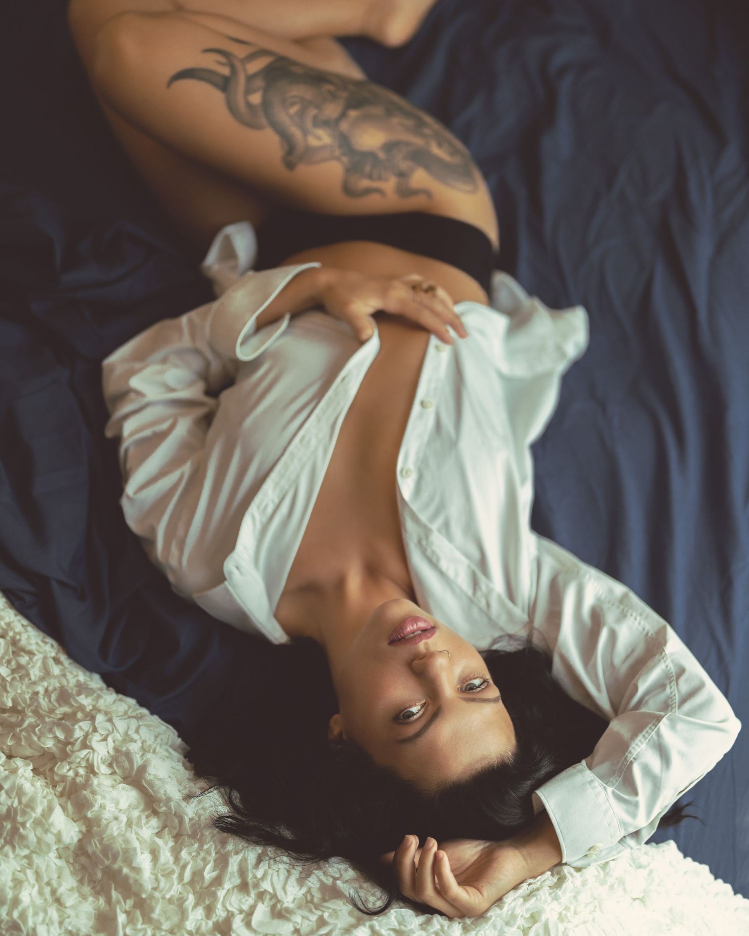 Tattooed woman lying on blue bedding wearing white shirt and black crop top in artistic pose.