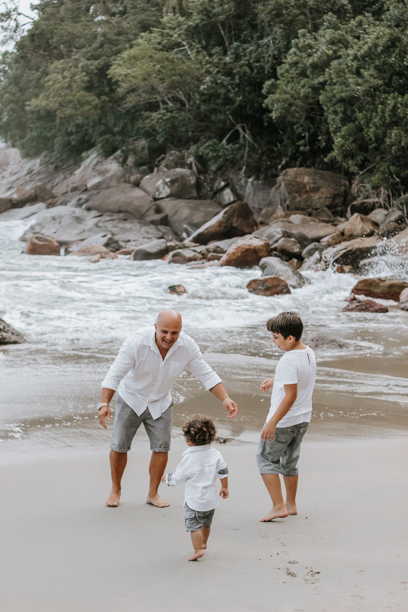 Pai e 2 filhos pequenos brincando, fundo de praia, mar e pedras, em Ubatuba.