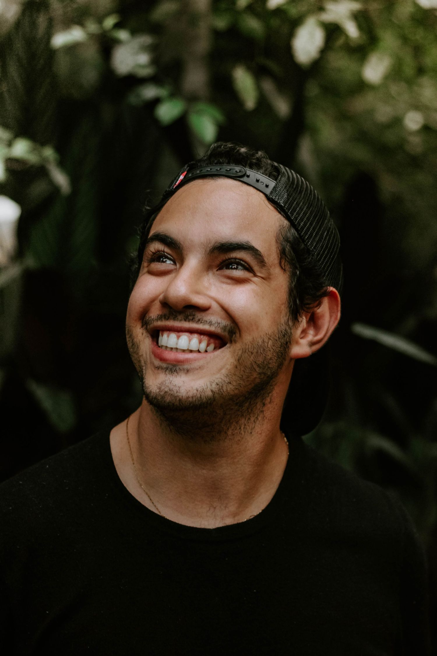 Retrato masculino em Ubatuba, homem jovem com boné preto e blusa preta de manga comprida, sorrindo ao ar livre, com natureza ao fundo.