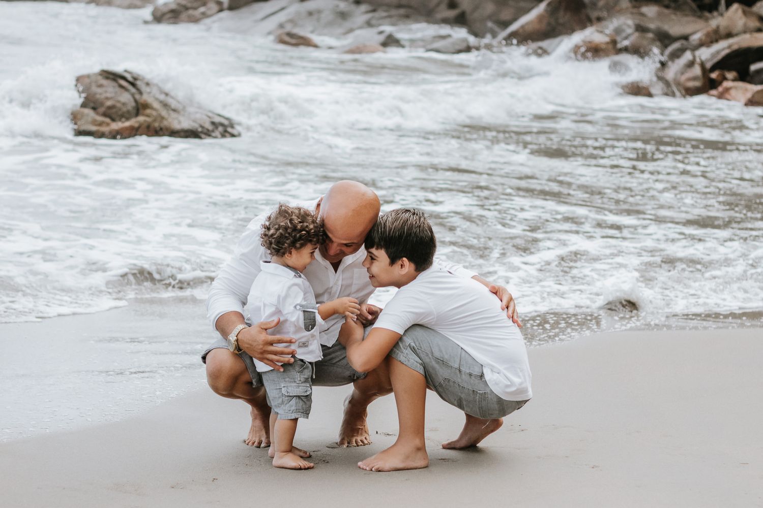 Pai e 2 filhos pequenos posando abaixados, fundo de praia, mar e pedras, em Ubatuba.