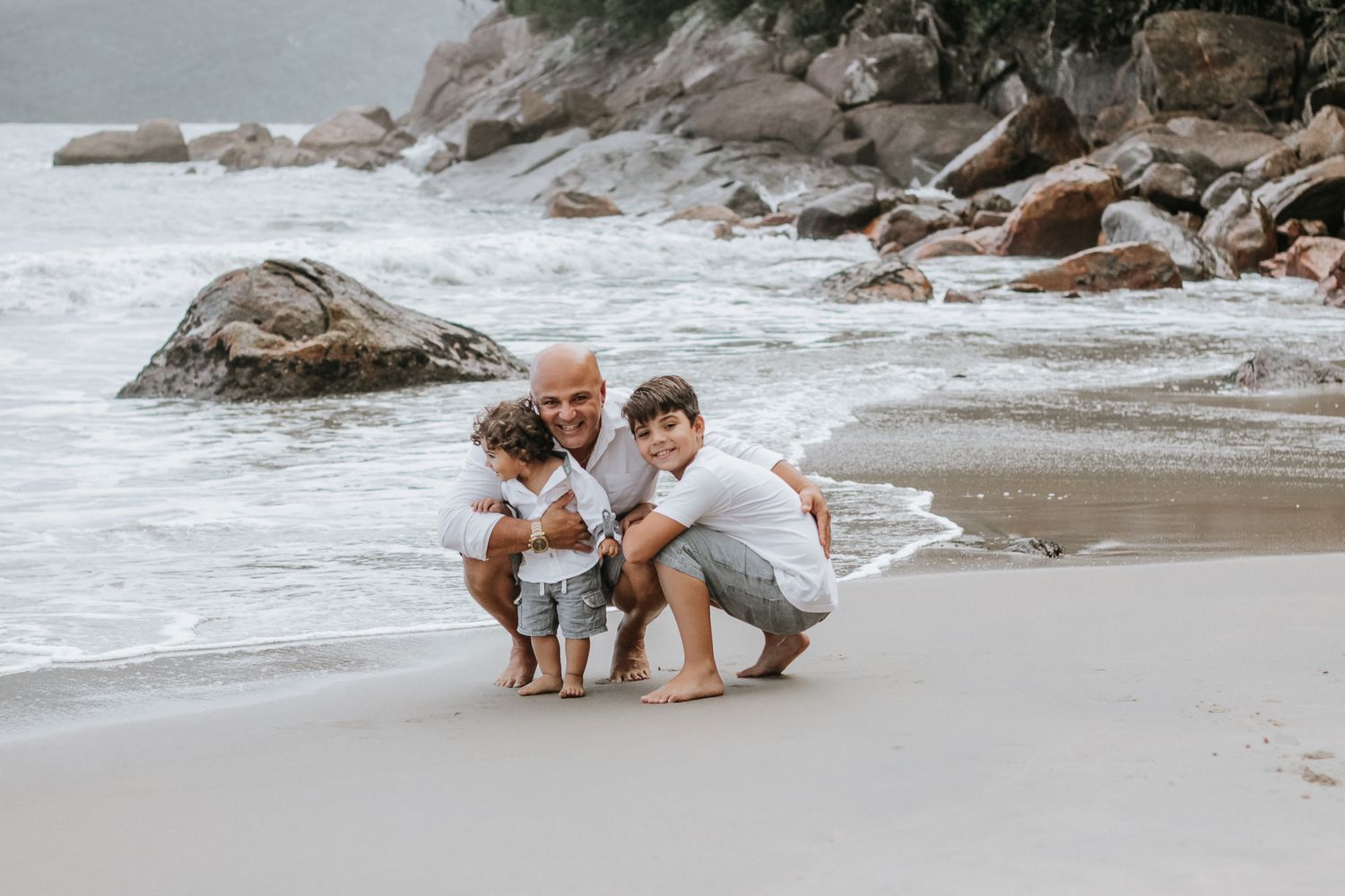 Pai e 2 filhos pequenos posando abaixados, fundo de praia, mar e pedras, em Ubatuba.