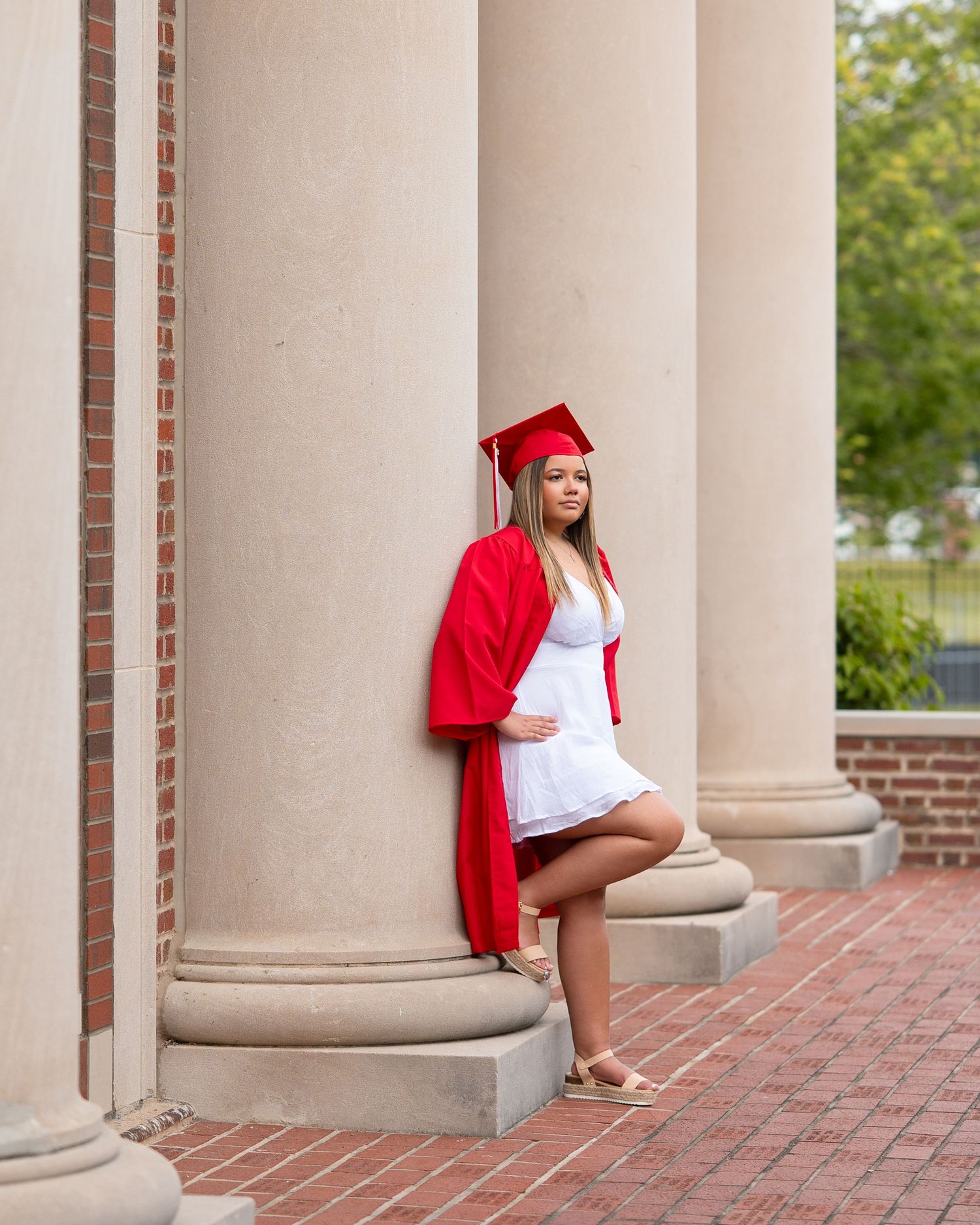 Pregnant graduate in red cap and gown leans against brick columns during outdoor senior portrait session.