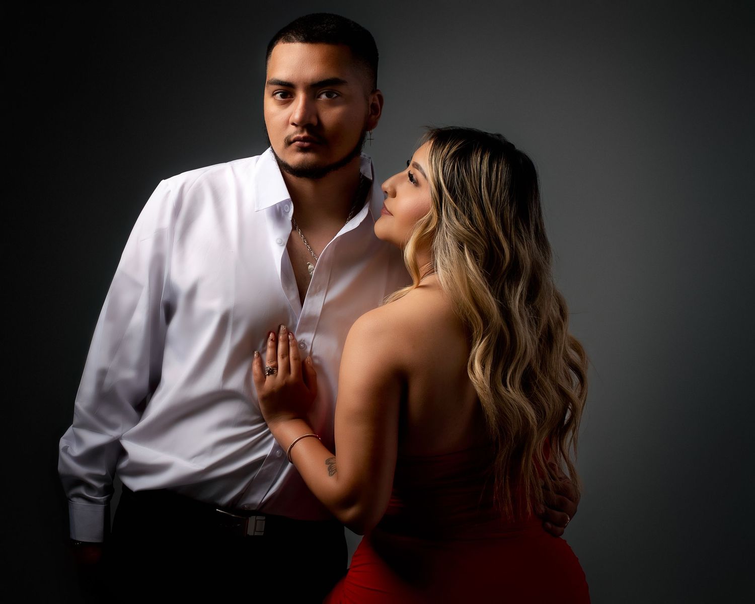 Couple in red dress and white shirt sharing intimate dramatic pose in dark studio setting.