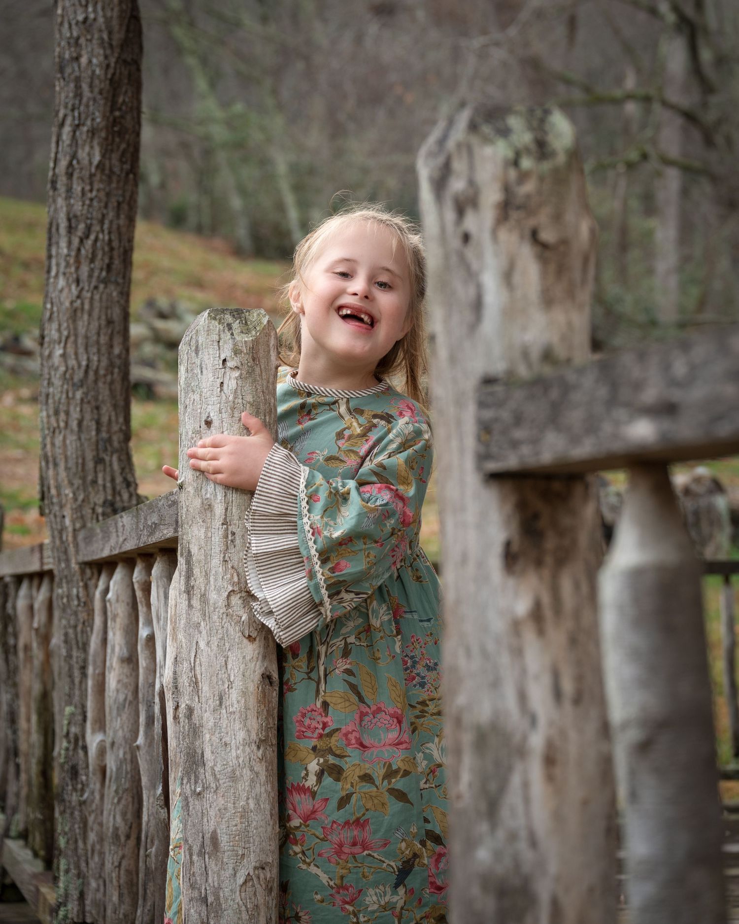 Happy little girl in floral green dress peeking and laughing from behind rustic wooden fence outdoors.