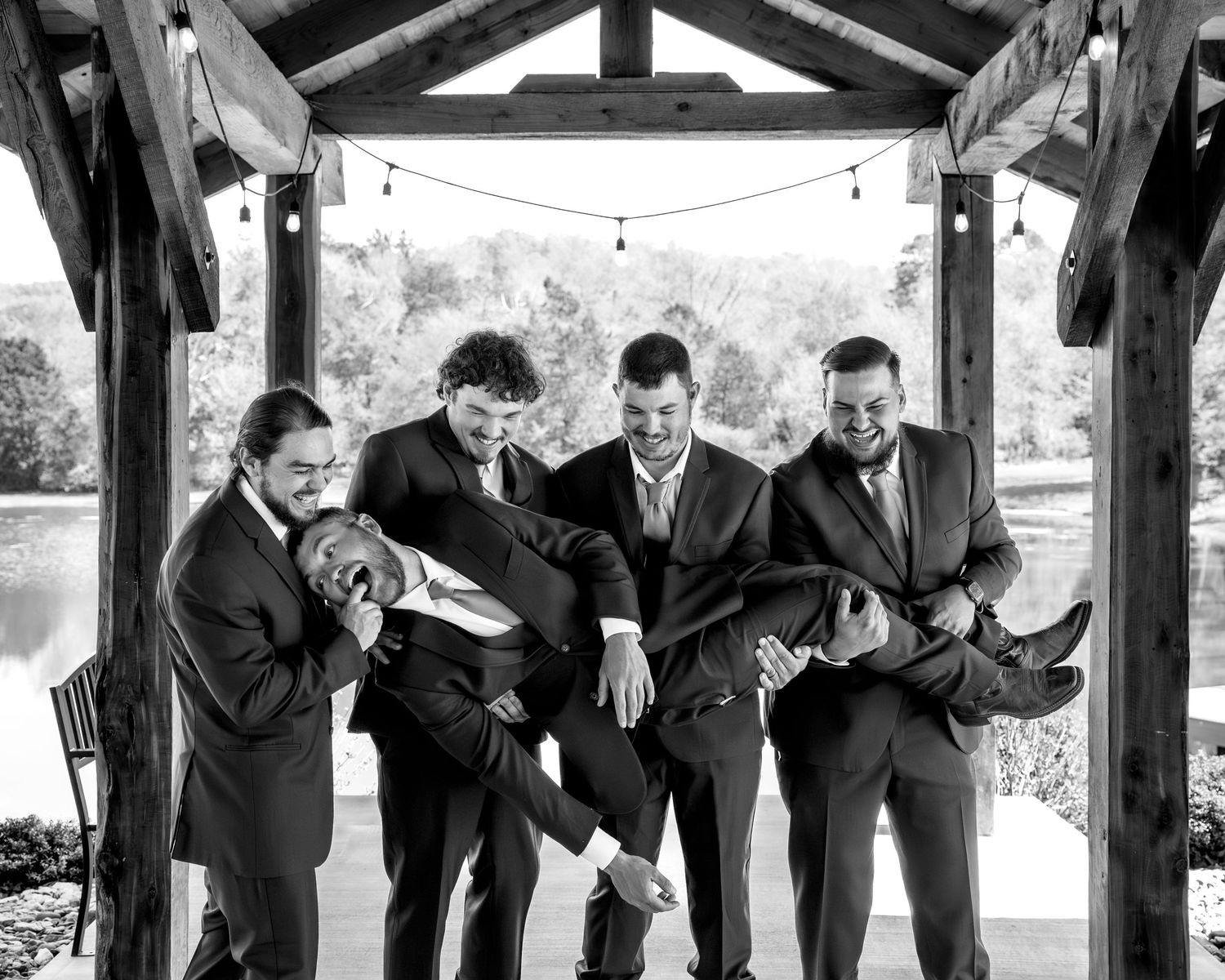 Black and white photo of groomsmen playfully lifting the groom under a rustic outdoor pavilion.