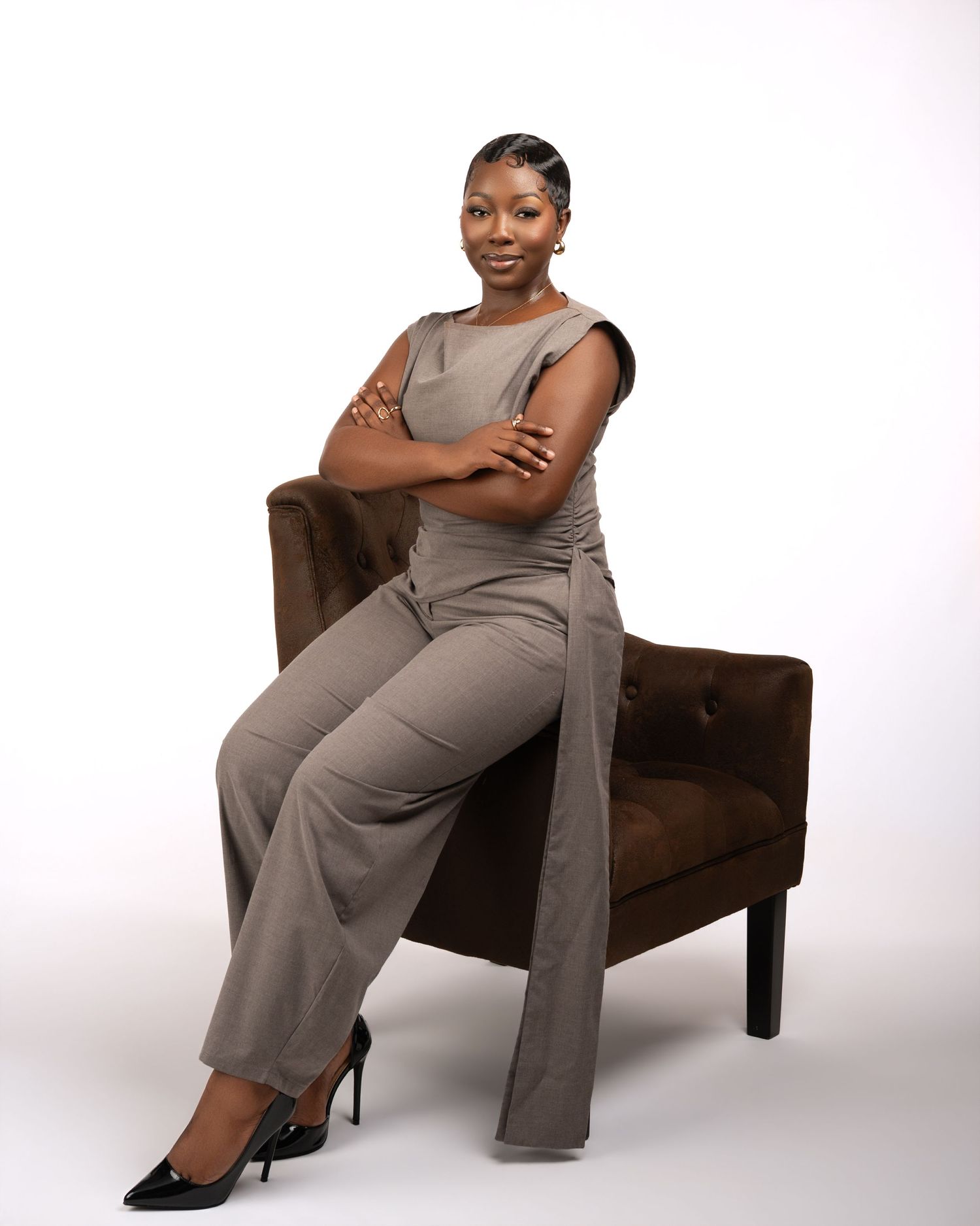 Professional woman in gray jumpsuit sitting confidently in brown chair with arms crossed against white background.