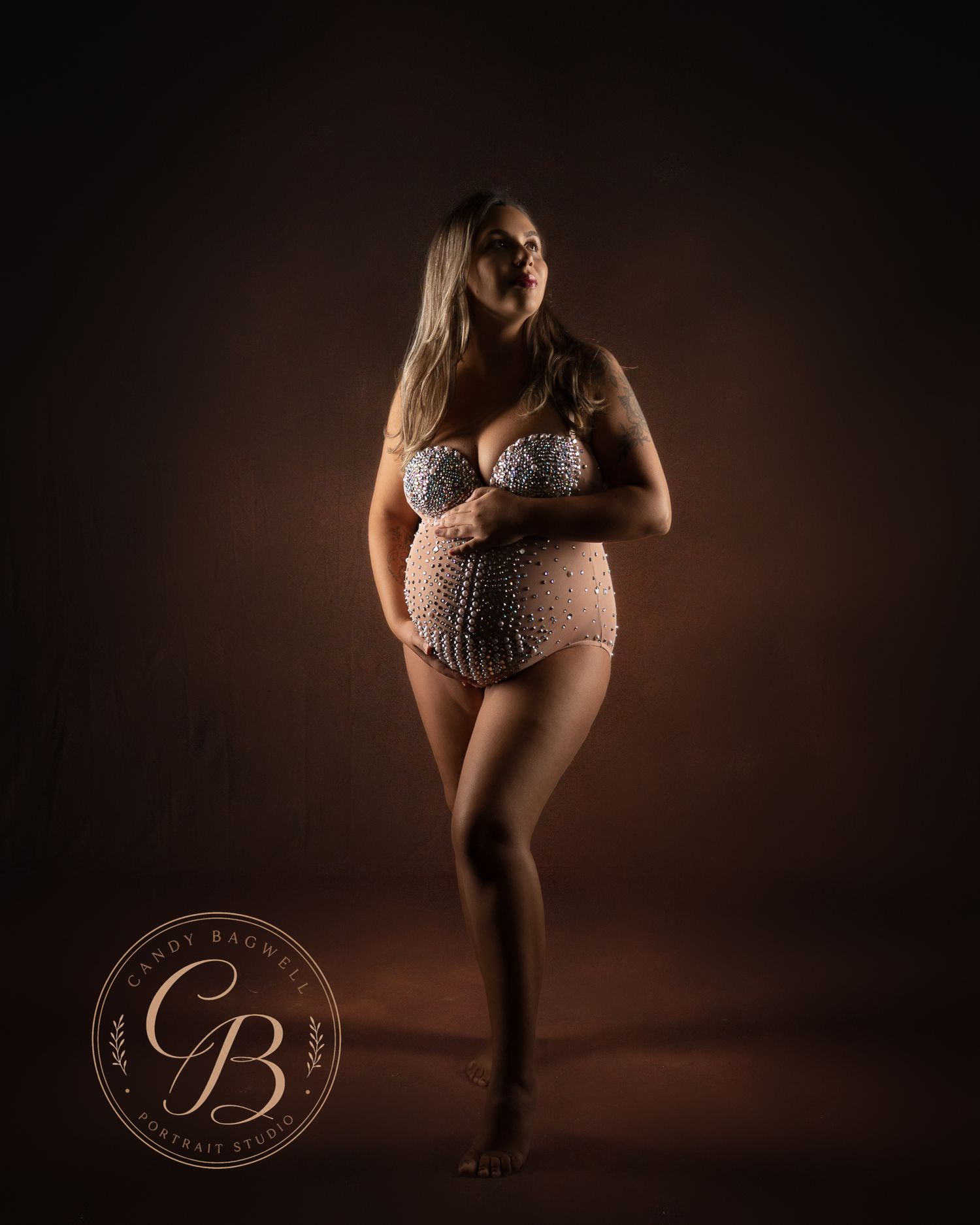 Pregnant woman in lace maternity lingerie poses gracefully against a warm brown backdrop in a professional studio portrait.
