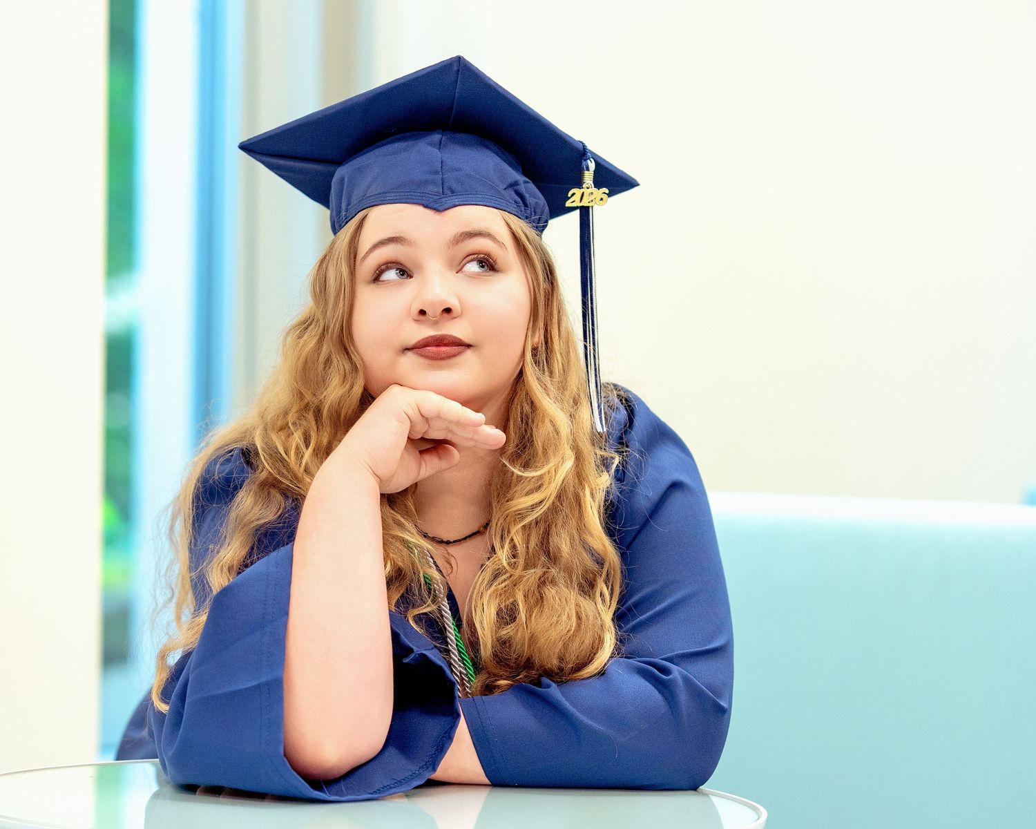 Female graduate in blue cap and gown resting chin on hand, looking upward thoughtfully indoors.