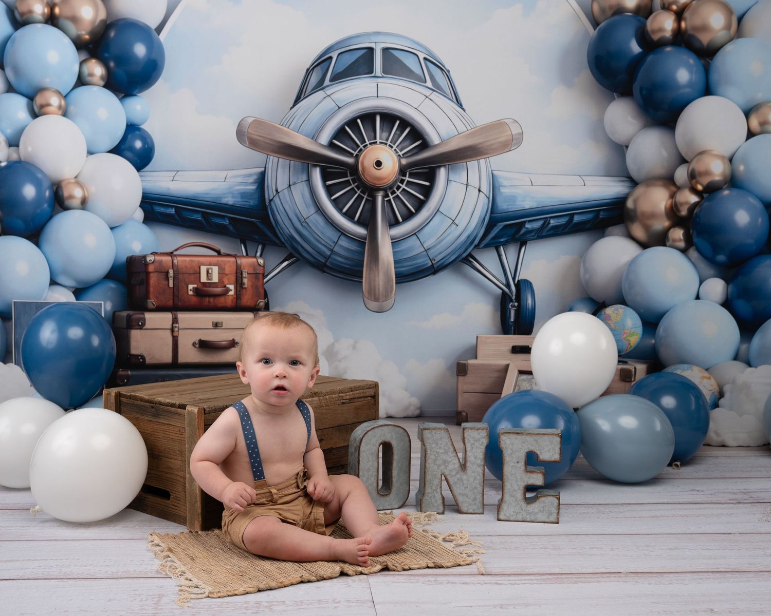 Baby boy in suspenders sits before airplane backdrop with blue balloons and ONE sign.