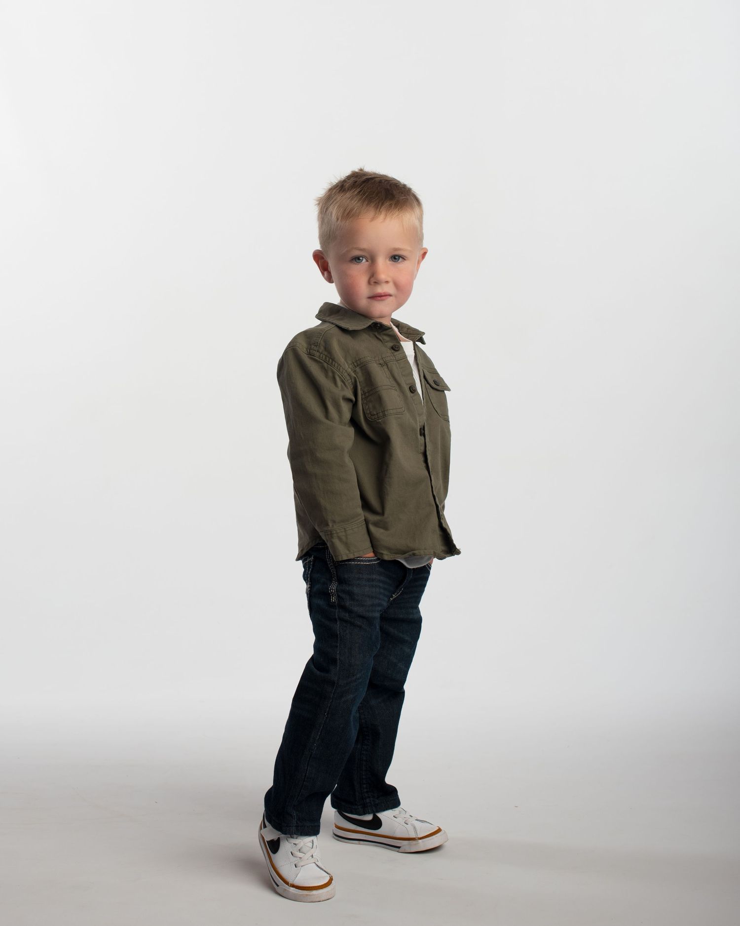 Smiling young blonde boy in olive jacket, jeans, and Nike sneakers posing on white studio background.