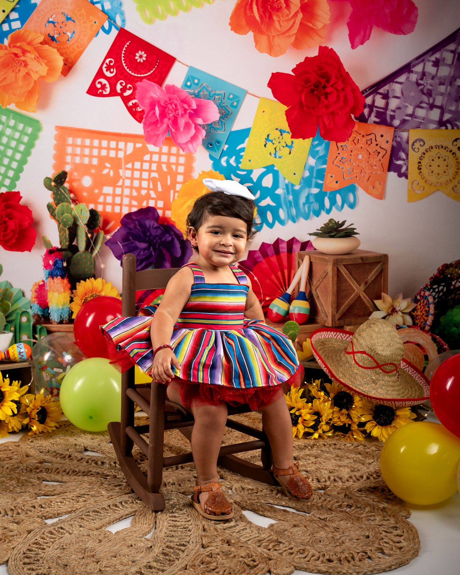 A smiling toddler in a colorful striped dress sits in a rocking chair with a festive fiesta-themed backdrop.